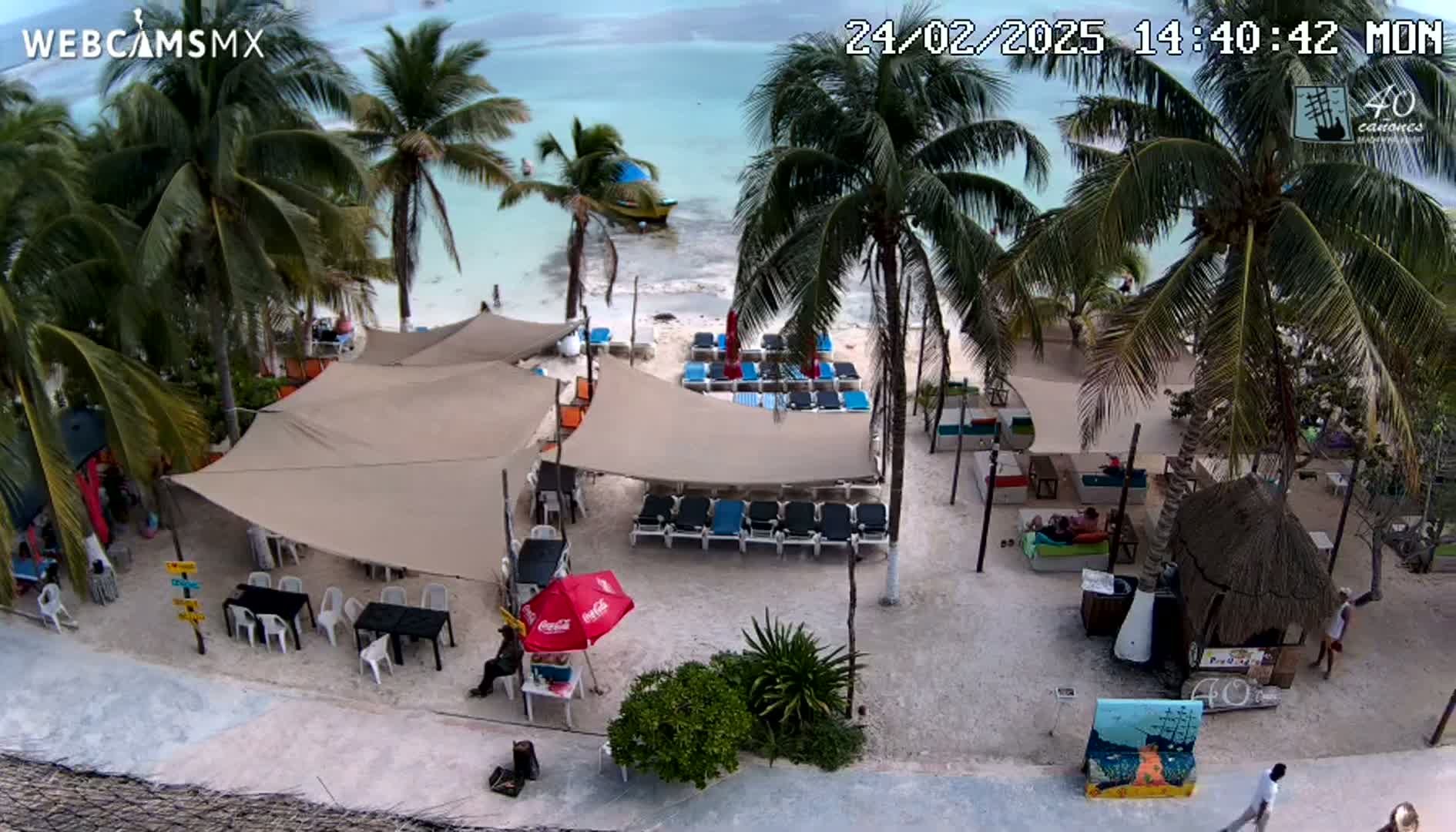 An overhead view of a beachside restaurant with lounge chairs and umbrellas under a partly sunny sky, overlooking a calm, light-blue ocean.