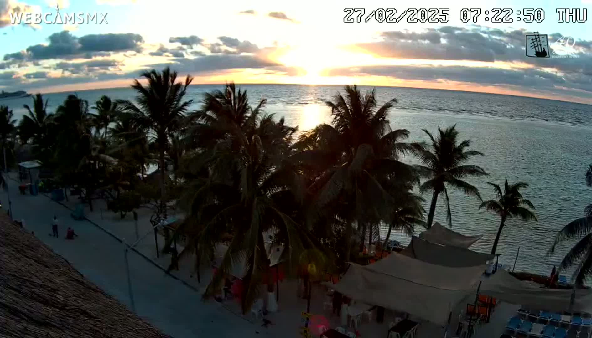 A sunset over a calm ocean is seen from an elevated vantage point, showing a beach with palm trees and outdoor seating areas.