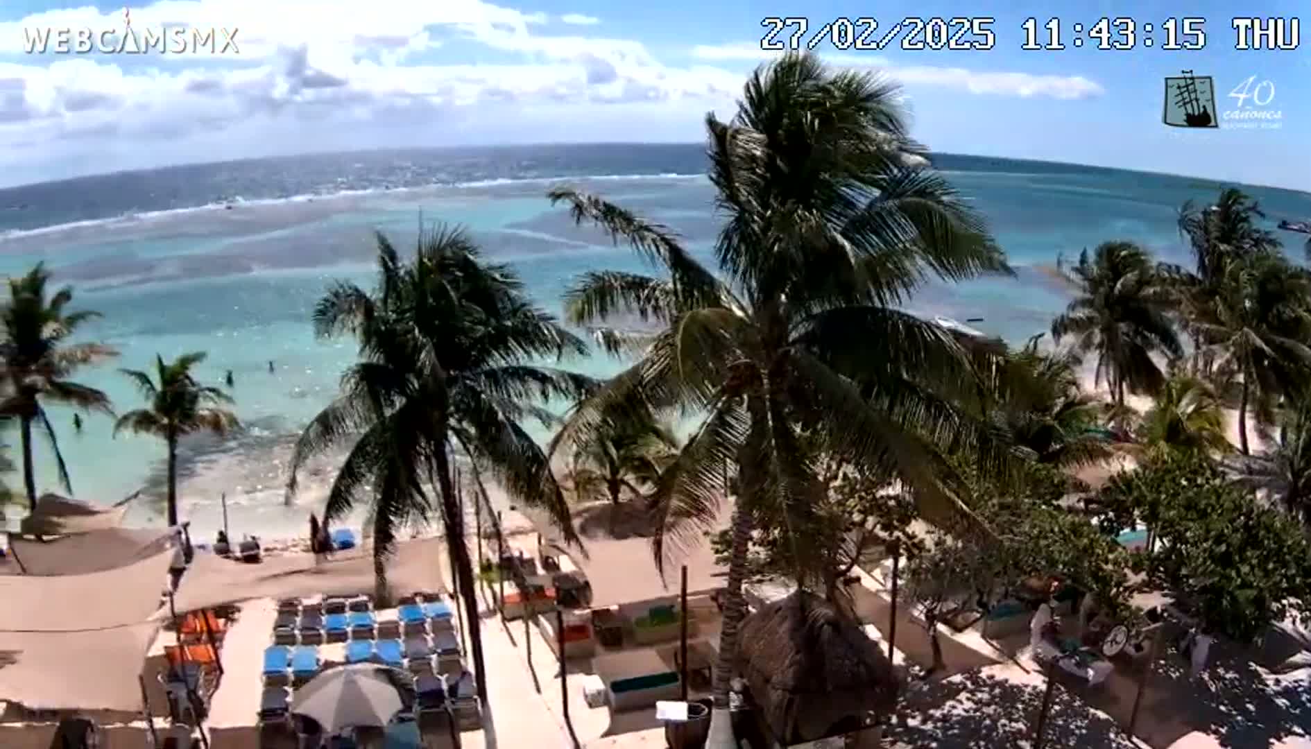 A partly sunny day shows a beach scene with palm trees, lounge chairs, and turquoise water extending to a distant horizon.
