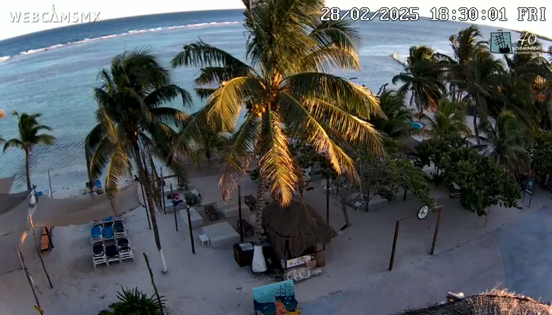 An aerial view shows a beach bar and lounge area on a sandy beach next to calm ocean water under a clear sky at sunset.