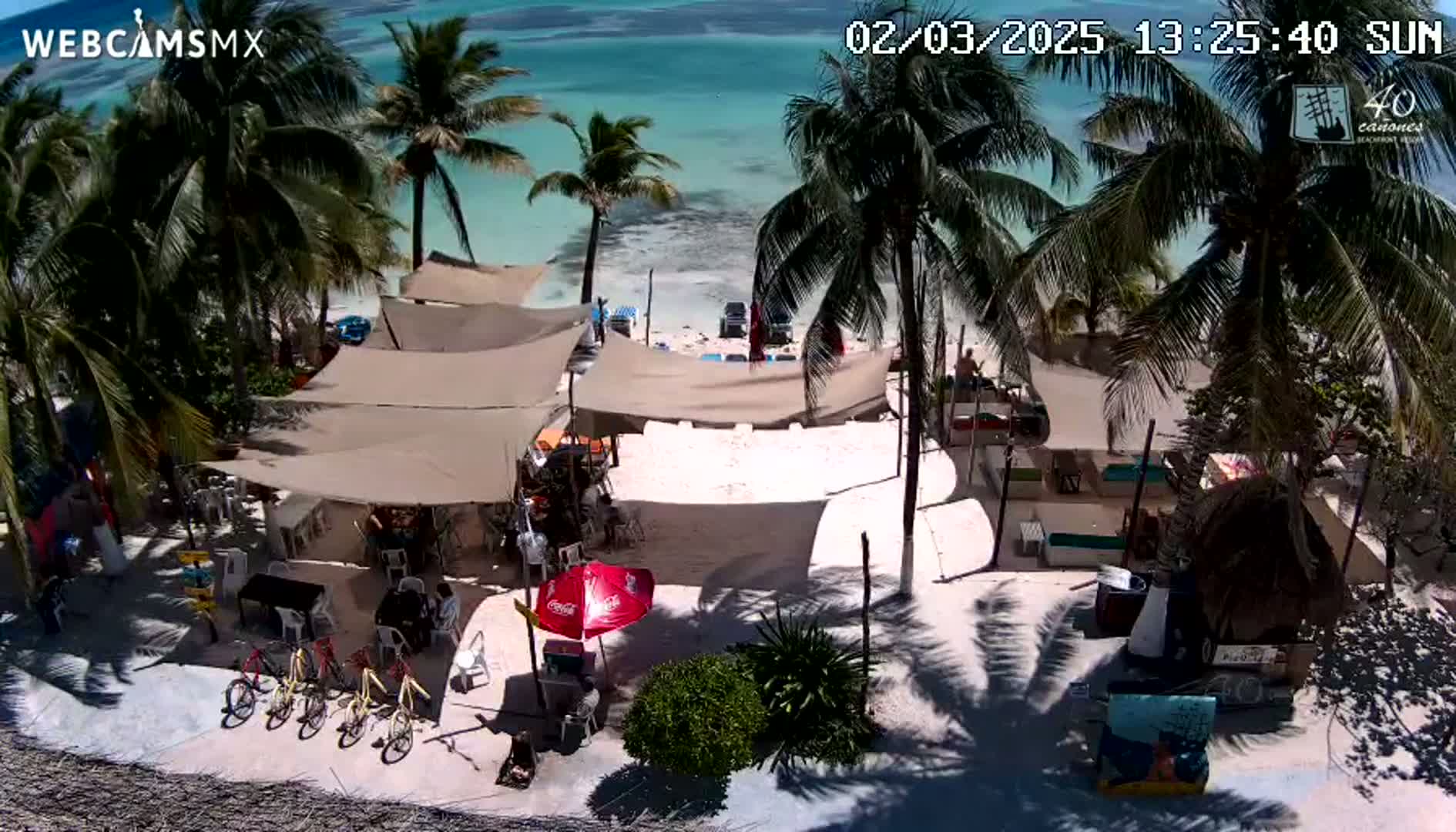 An overhead view of a beach bar with several umbrellas and tables on a sunny day, next to a clear, turquoise ocean and surrounded by palm trees.