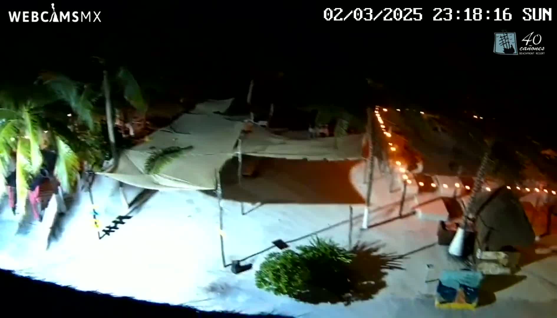 An aerial nighttime view shows a sandy area with several canopies, string lights, and palm trees under a clear, dark sky.