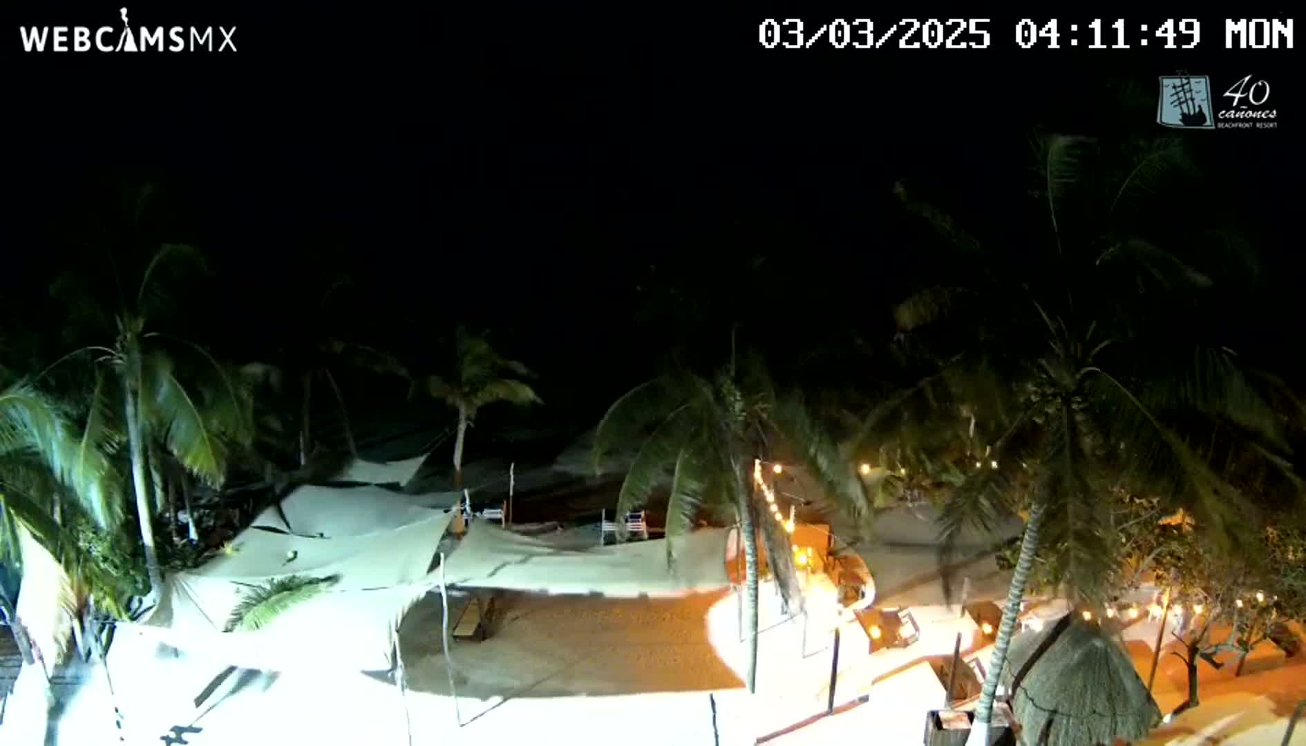 A nighttime, high-angle view of a beachside resort with palm trees, illuminated white canopies, and string lights, under a clear, dark sky.