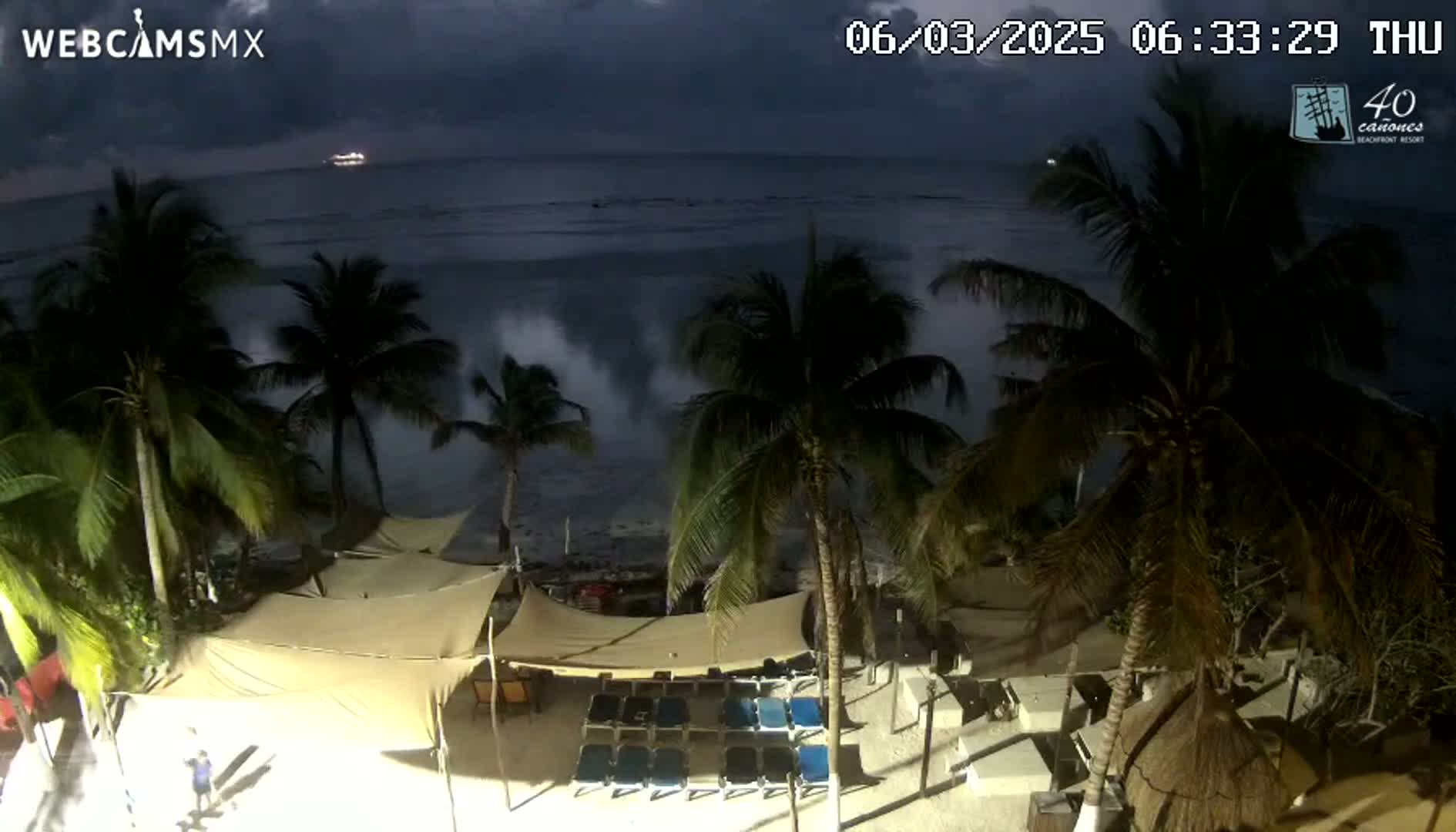 Under a dark, cloudy night sky, large waves crash against a beach lined with palm trees and beachside lounge chairs under canopies.