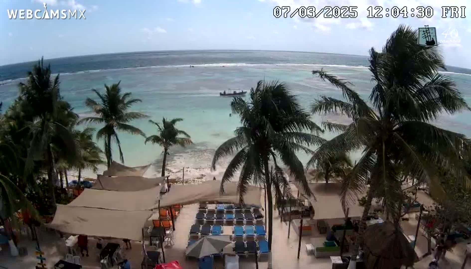 A beachside restaurant with lounge chairs and umbrellas overlooks a calm, light-blue ocean under a mostly sunny sky with a gentle breeze blowing palm trees.