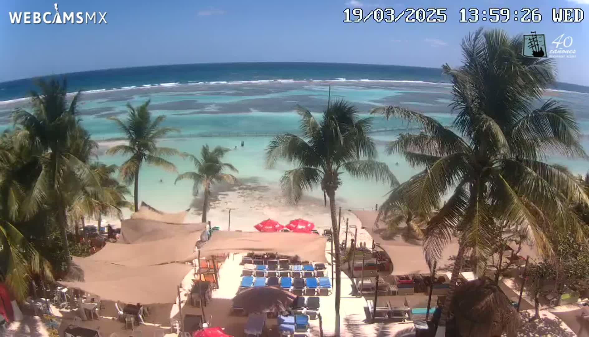A beachside restaurant with lounge chairs and umbrellas overlooks a calm, turquoise ocean under a sunny sky.