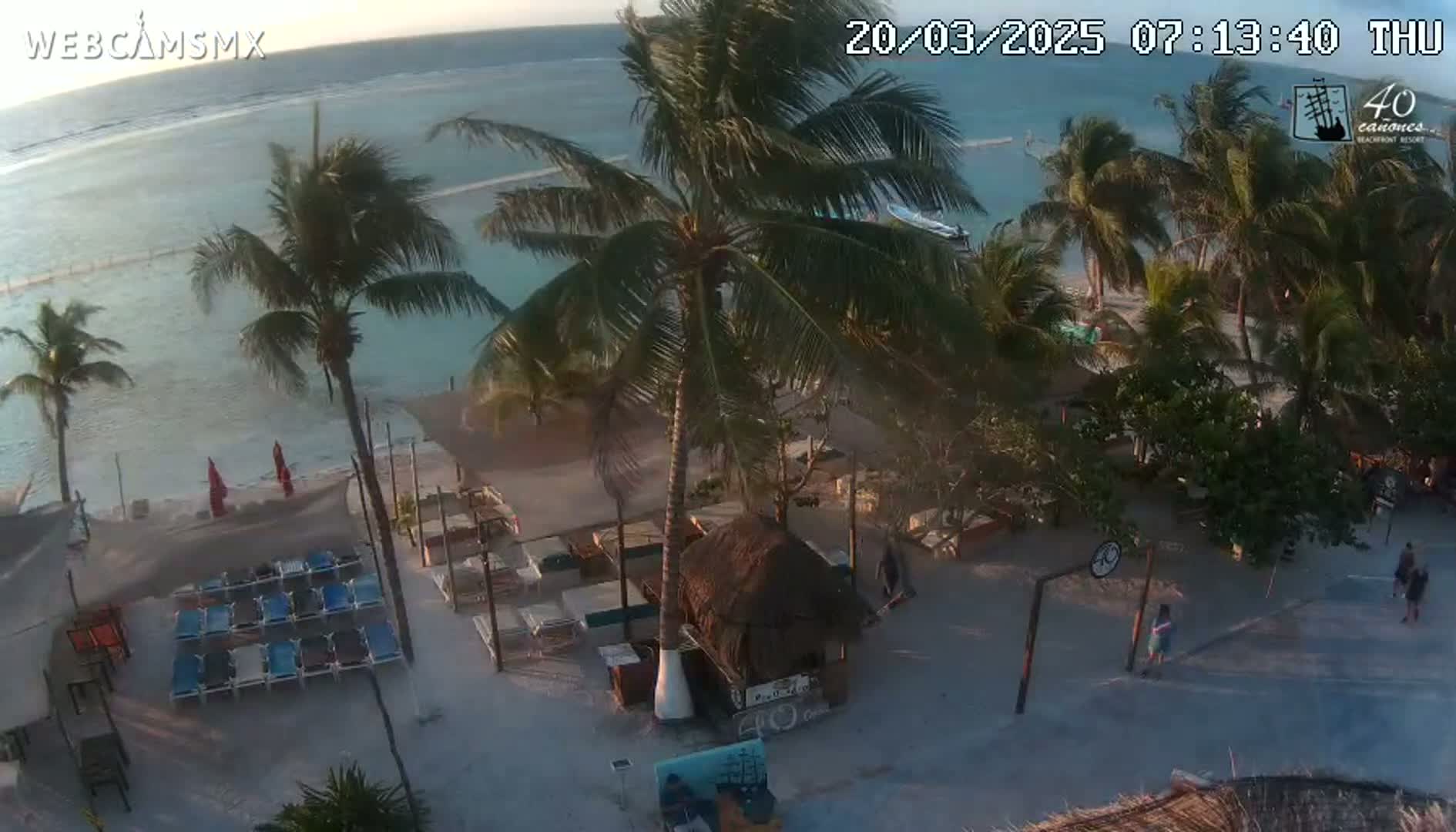 An aerial view of a beachside area shows calm, light-blue ocean water, palm trees, beach chairs, a small thatched-roof structure, and a few people near a basketball hoop under a mostly clear sky.
