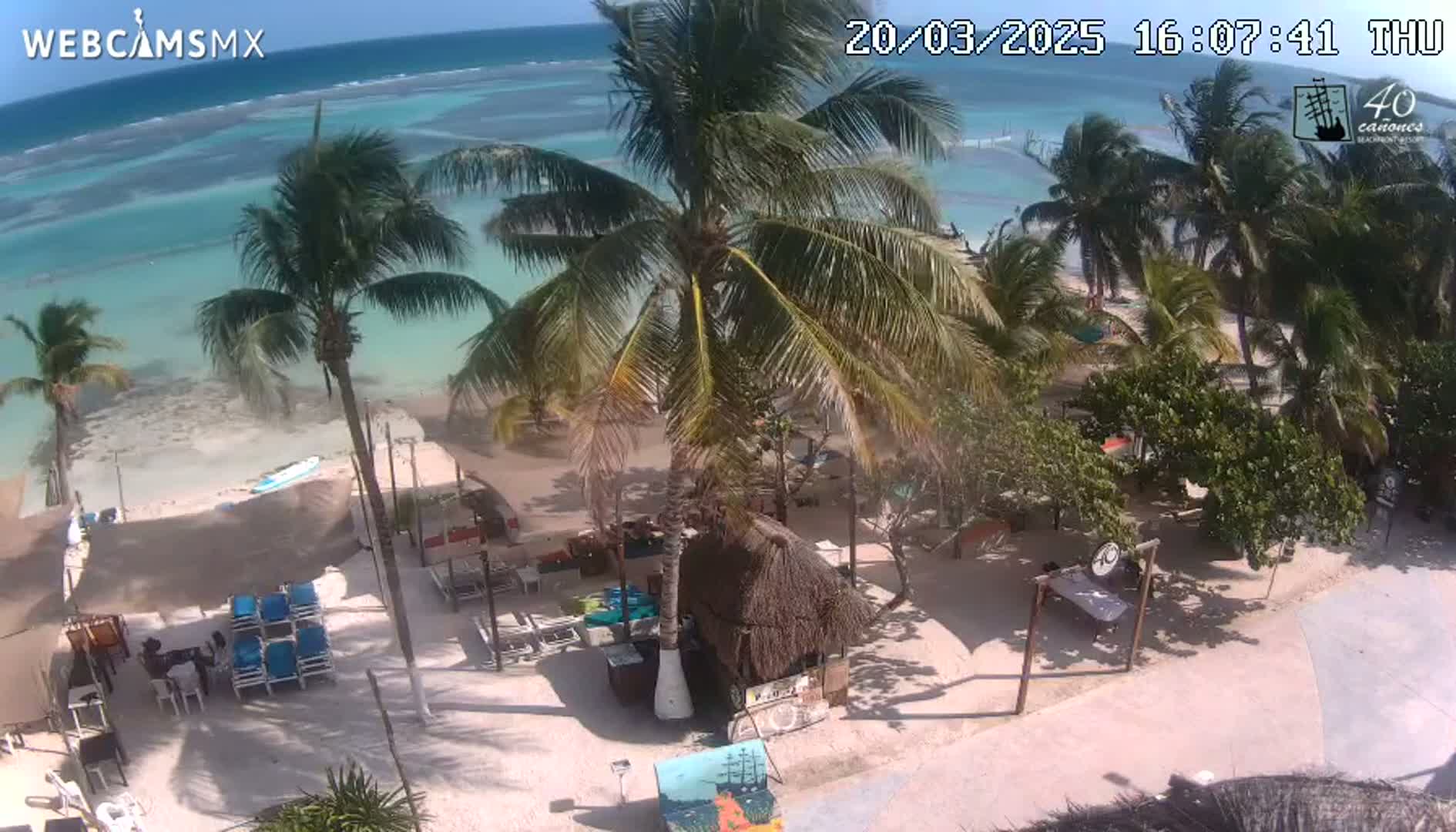 An overhead view shows a beachside area with palm trees, beach chairs, and small structures, under sunny skies and a clear, light-blue ocean.
