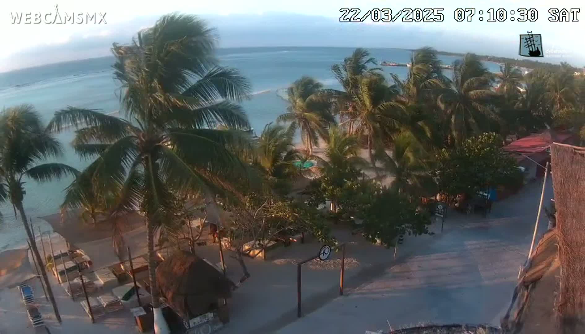 An aerial view of a tropical beach scene with palm trees, calm ocean water, and a small village under a partly sunny sky with a gentle breeze.