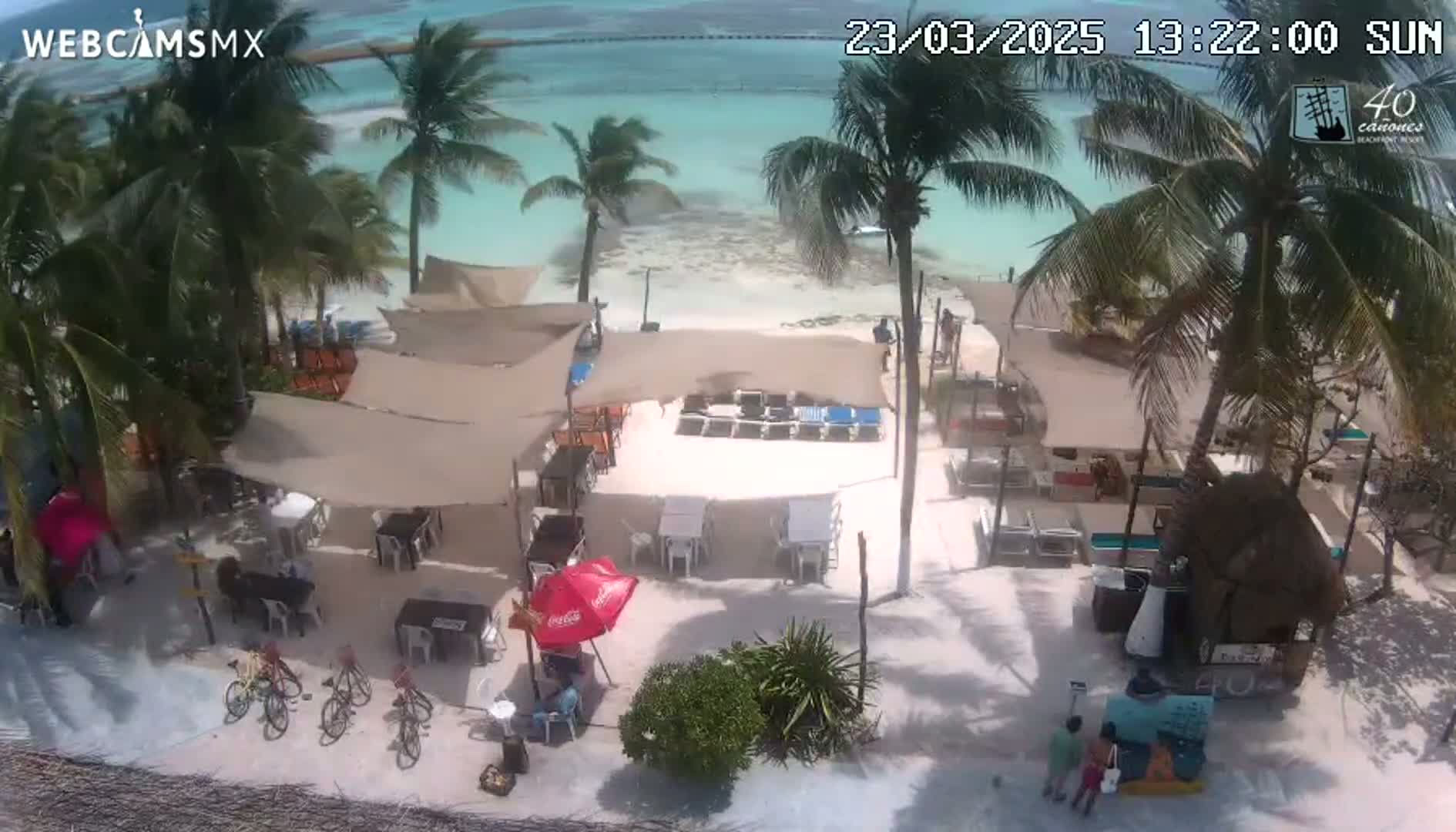 An aerial view shows a beachside restaurant with shaded seating areas, lounge chairs, and several bicycles parked on a sandy area under a sunny sky.