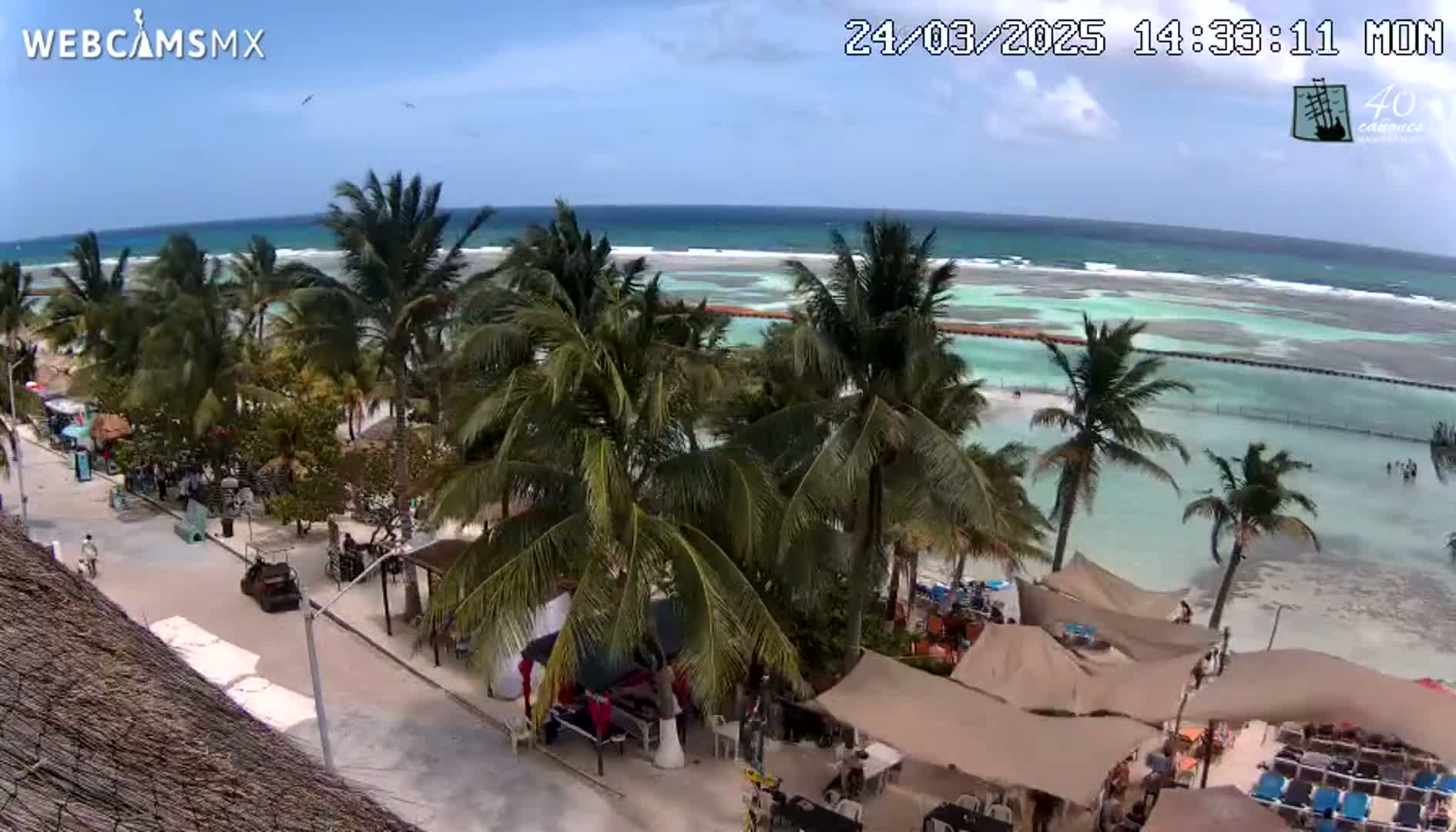 A beachside street lined with palm trees and beach umbrellas, overlooking a calm, turquoise ocean under a mostly sunny sky.