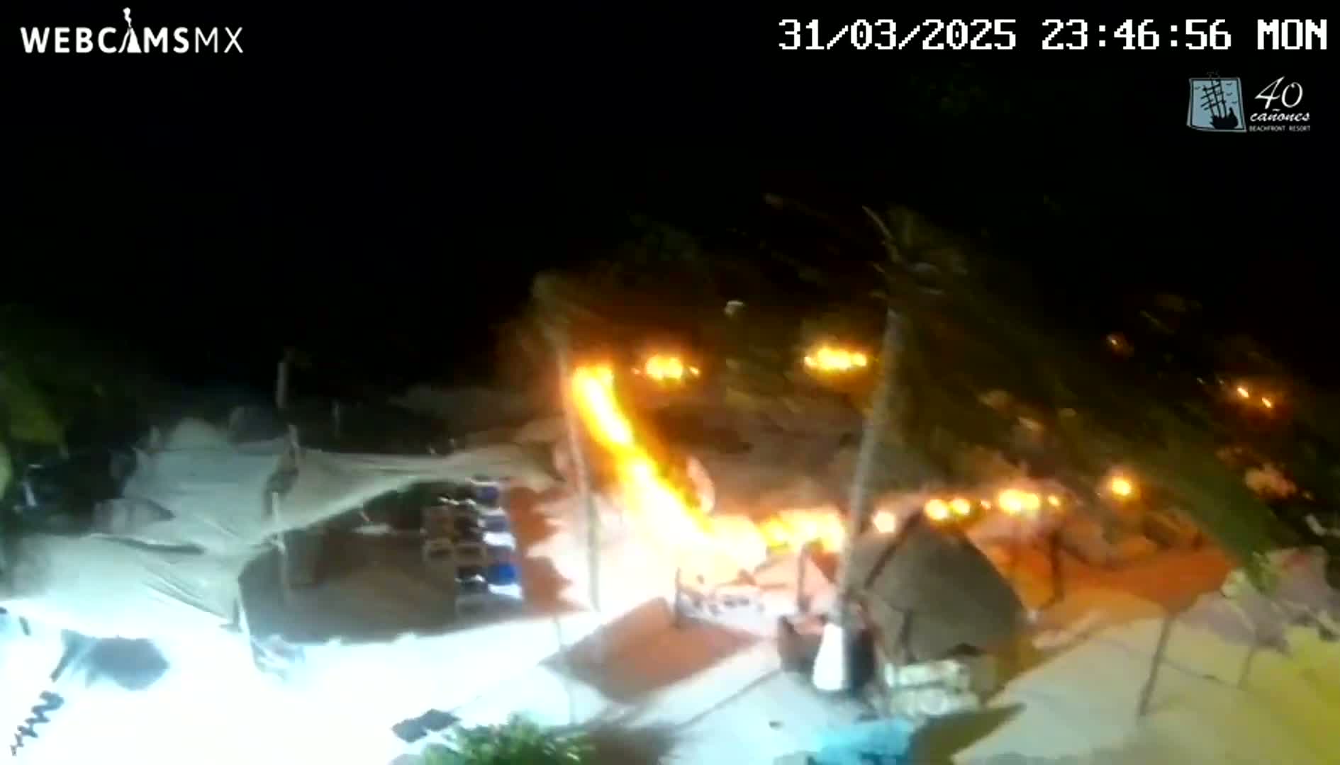 A nighttime beach scene shows a long, bright flame shooting upward near several small structures, surrounded by many smaller, glowing lights.