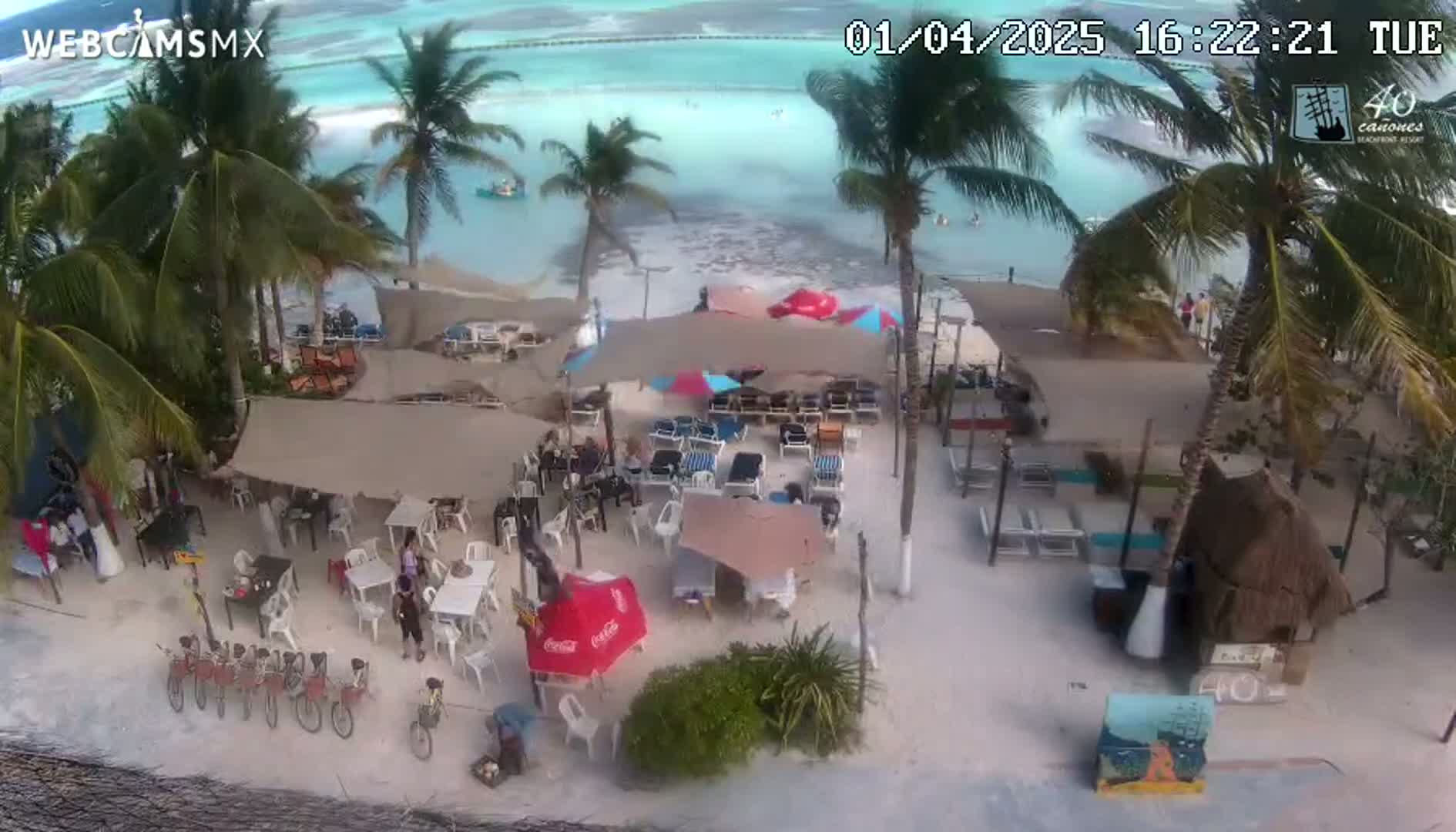 An aerial view of a beachside restaurant with numerous tables and chairs under umbrellas, several bicycles parked nearby, and palm trees surrounding a sandy beach with calm, turquoise water visible in the background.