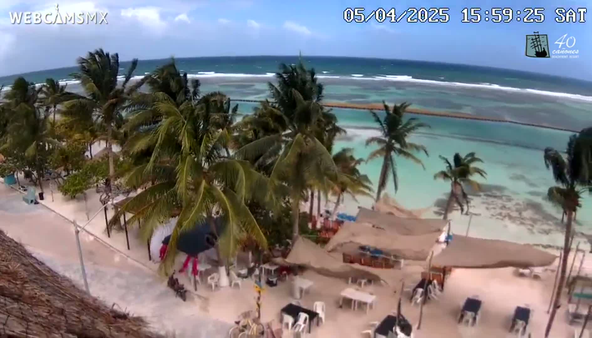 A beachside restaurant area with palm trees is shown, overlooking a turquoise ocean with moderate waves under a partly cloudy sky.