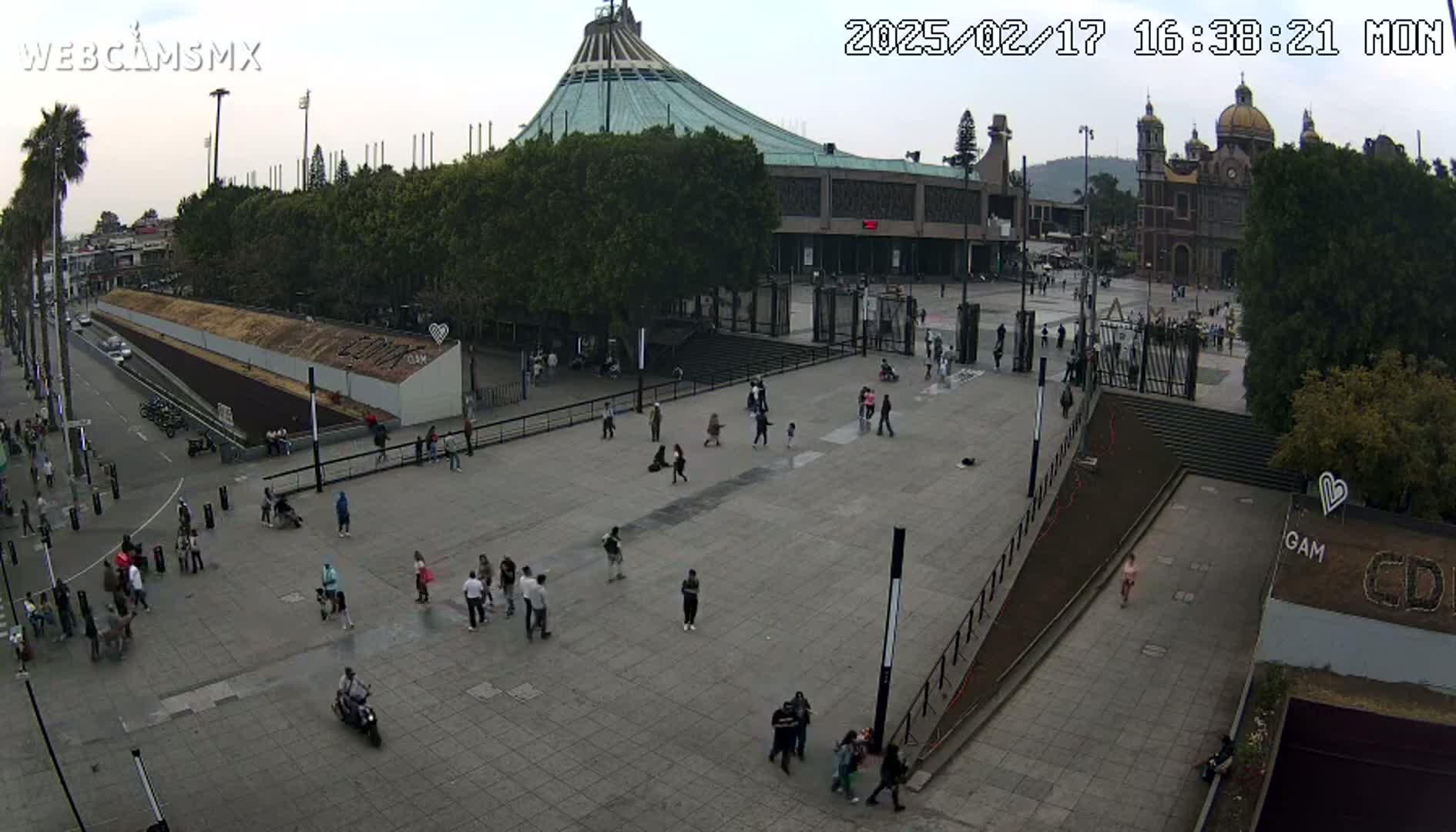 A large plaza with scattered people, a large modern cathedral, and a smaller basilica-style church in the background under an overcast sky.