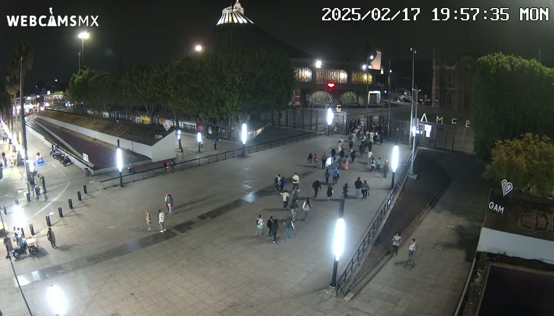 A nighttime, outdoor view of a plaza with numerous people walking, near a large building with a dome.