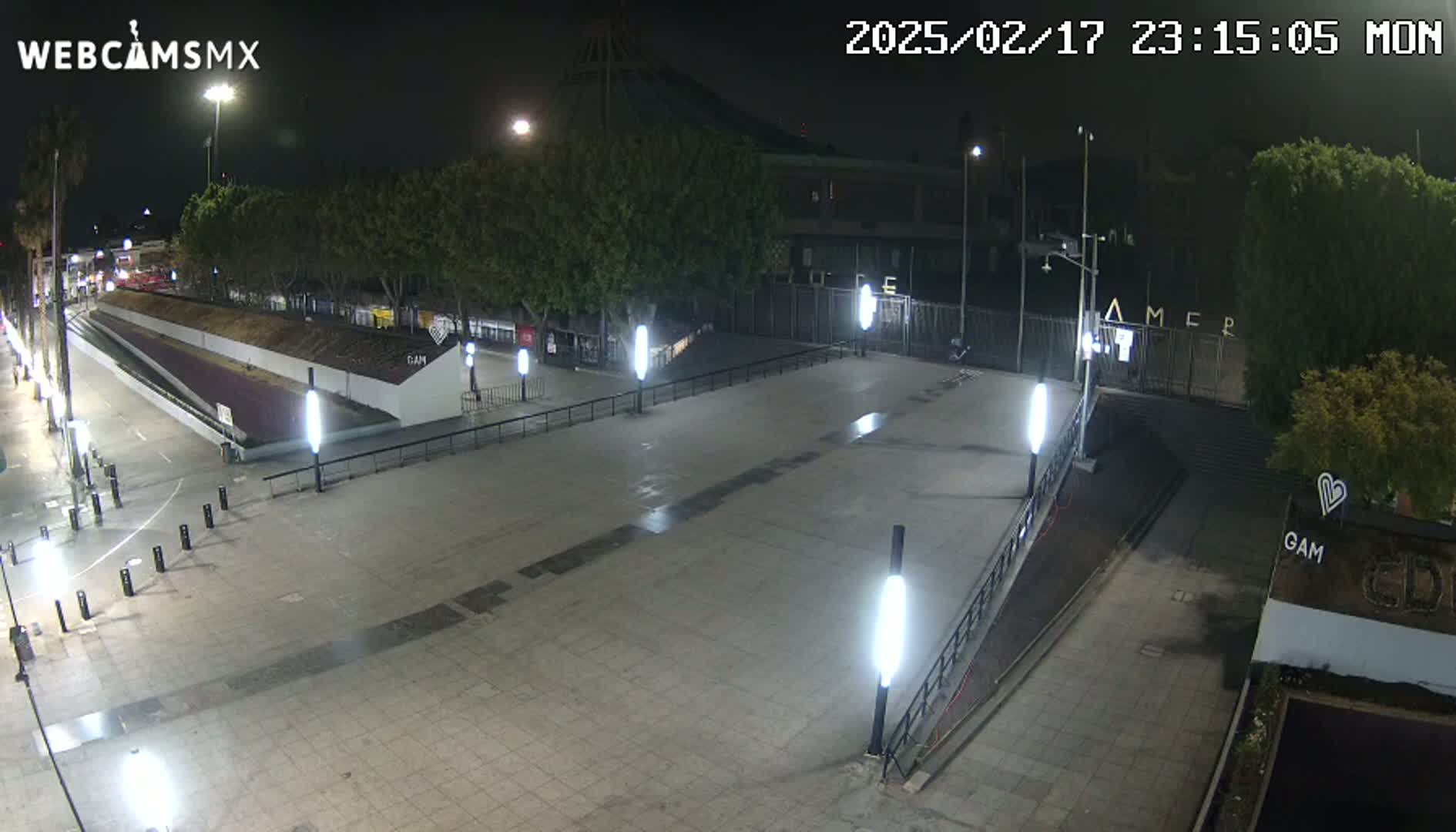 A nighttime, paved plaza with several tall lights is mostly empty except for some small objects and a few trees in the background.