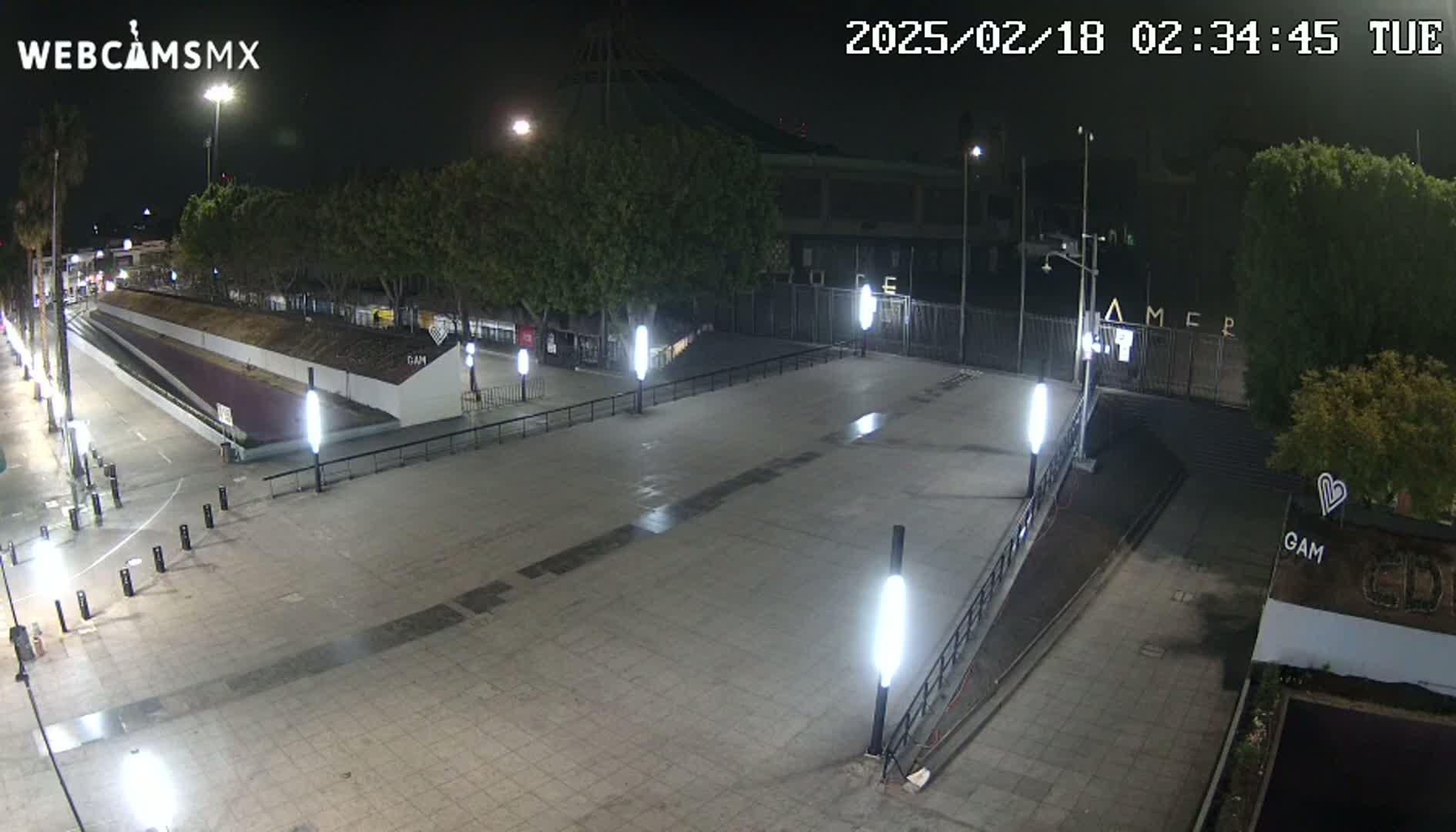 A nighttime, paved plaza area with several tall lights, trees, and a gated entrance to a building in the background.