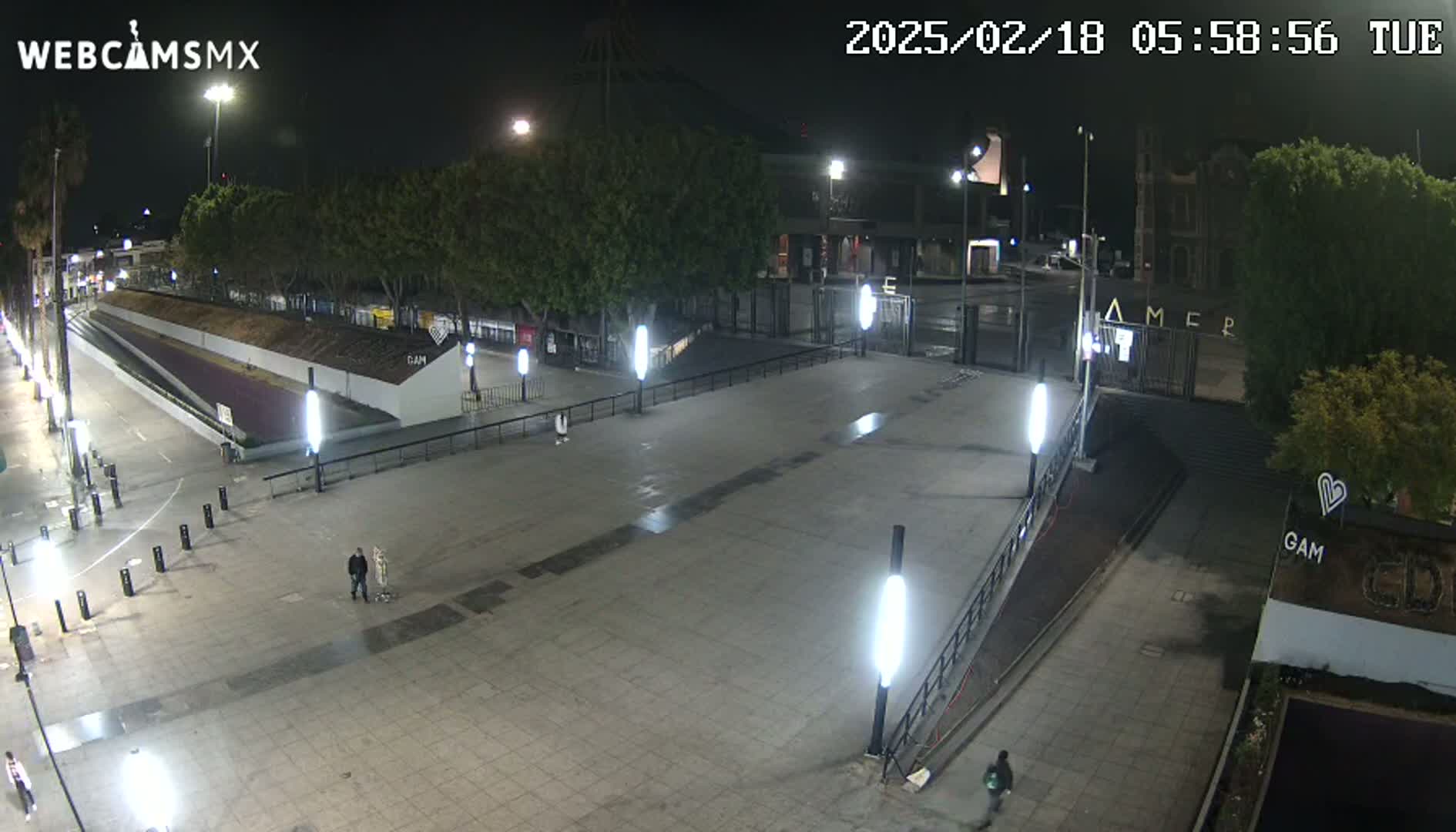 A mostly empty plaza at night, illuminated by bright lights, with a few people visible and some trees in the background.