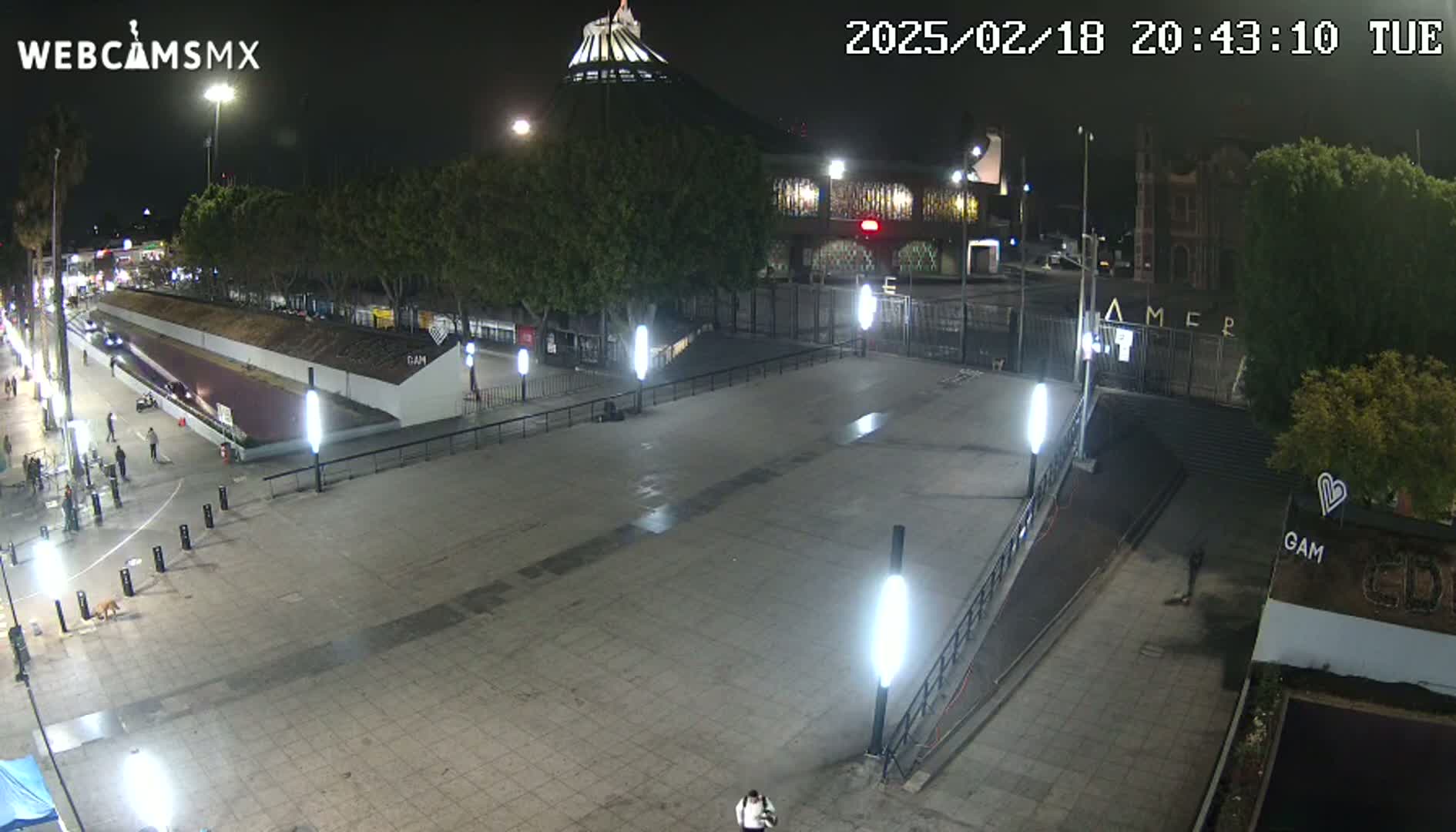 A mostly empty plaza at night, illuminated by many lights, is situated in front of a large building and gated entrance.
