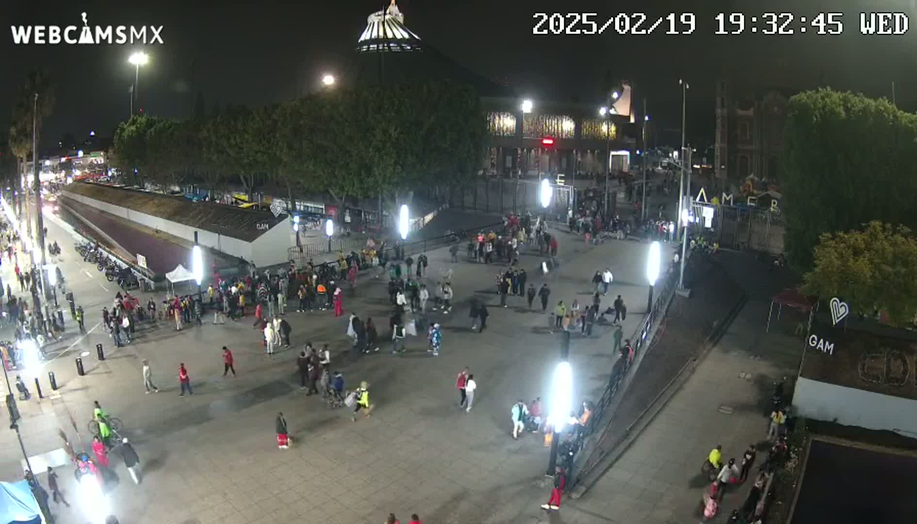 A nighttime aerial view of a plaza with numerous people gathered, illuminated by streetlights, near a large building under a clear sky.