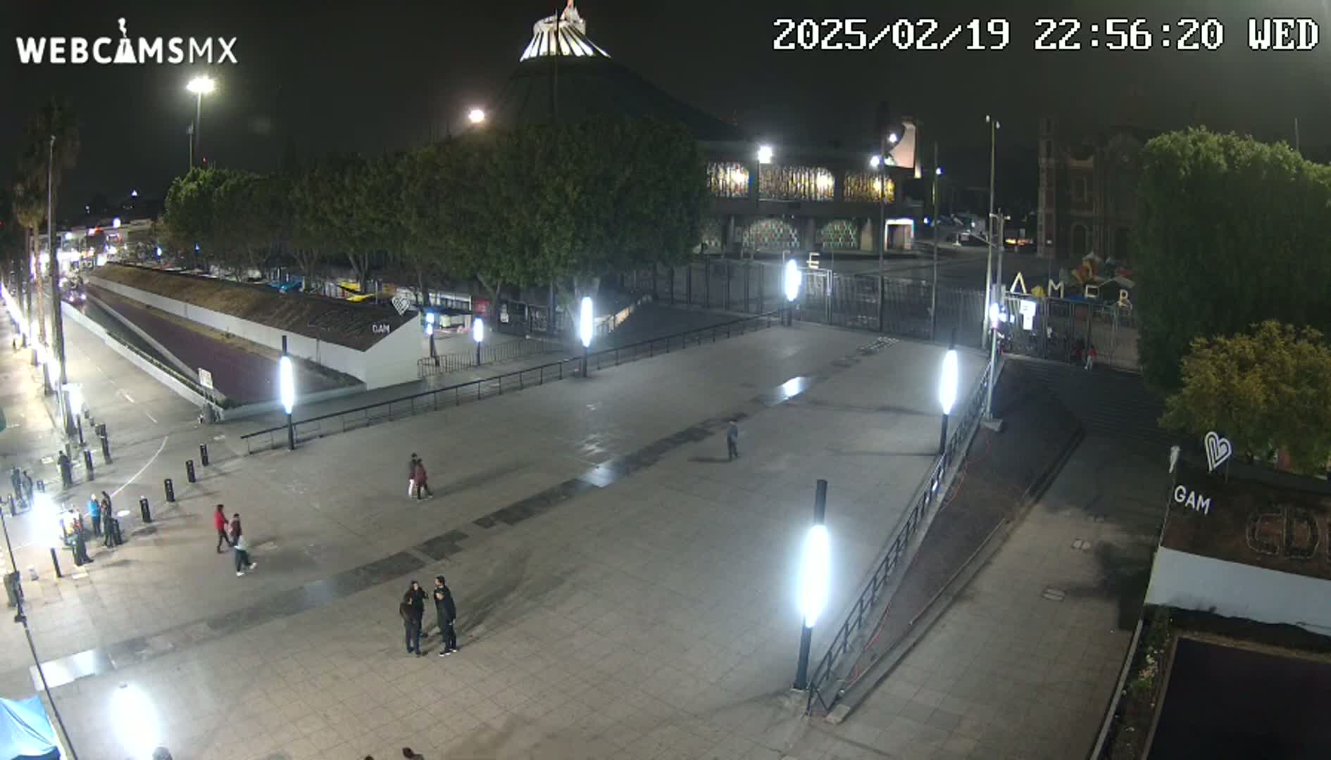 A nighttime, high-angle view of a plaza with several people walking around, illuminated by bright lights, in front of a large building.