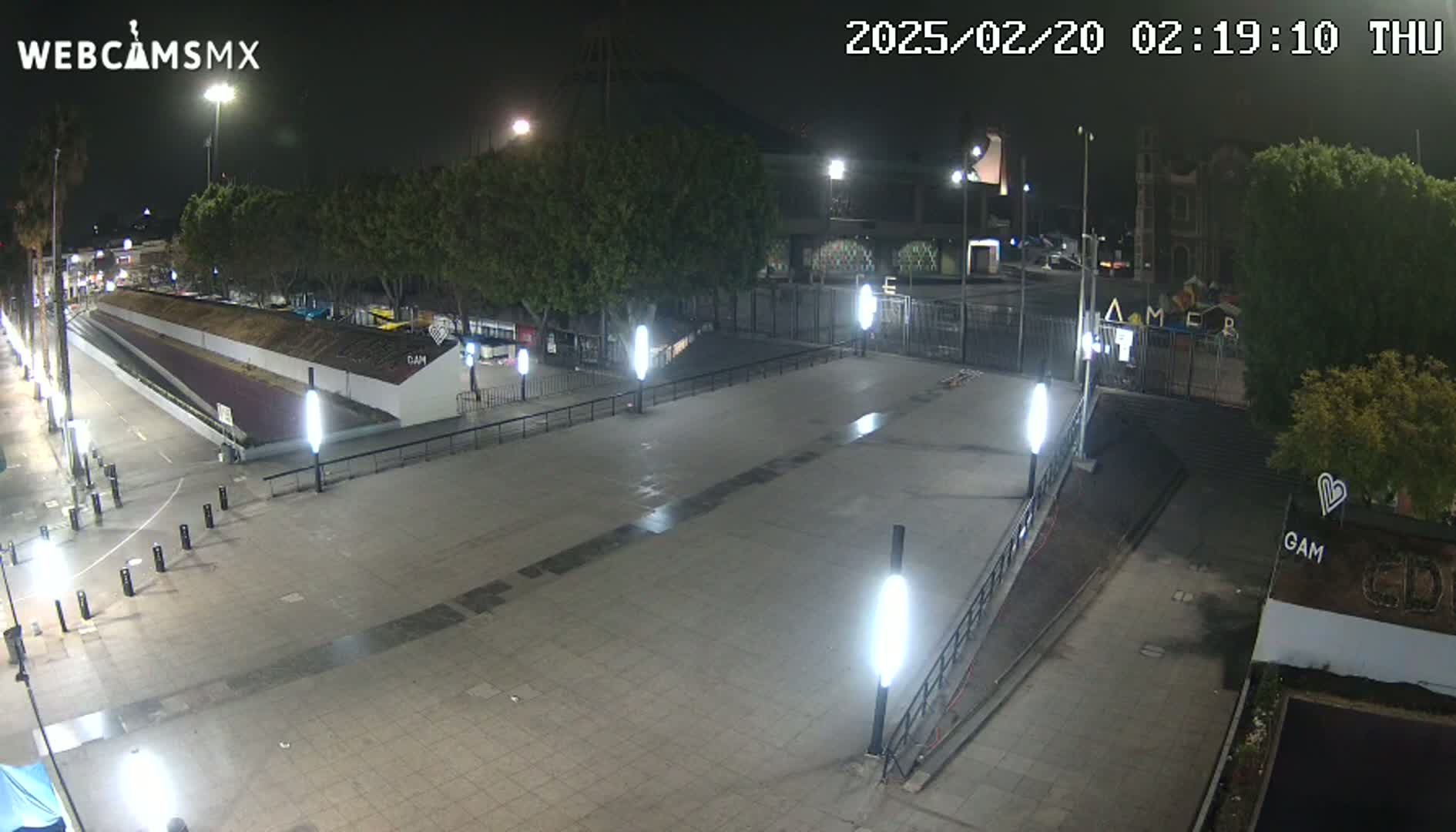 A nighttime view of an empty plaza with several lit lampposts, bordered by trees and buildings.