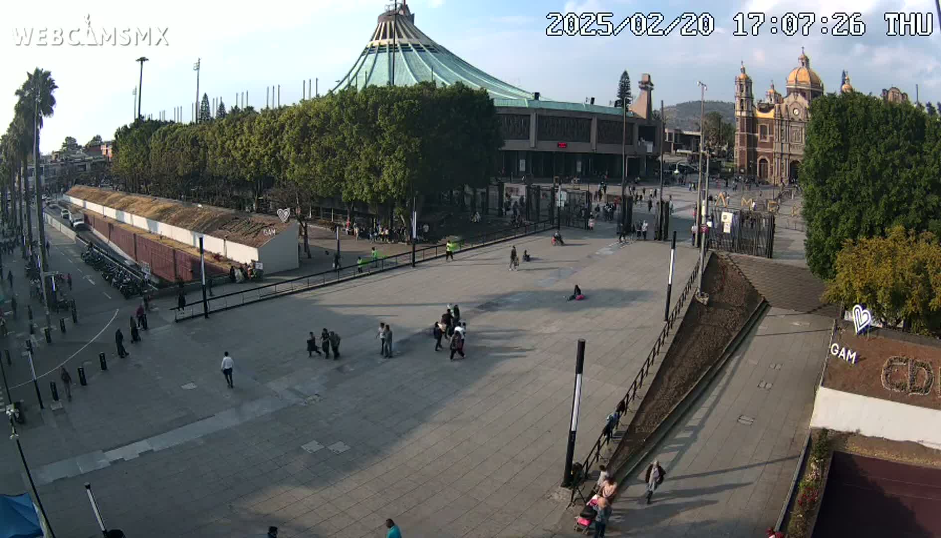 A large plaza with several people walking around, in front of a basilica and a large modern building, under a mostly sunny sky.