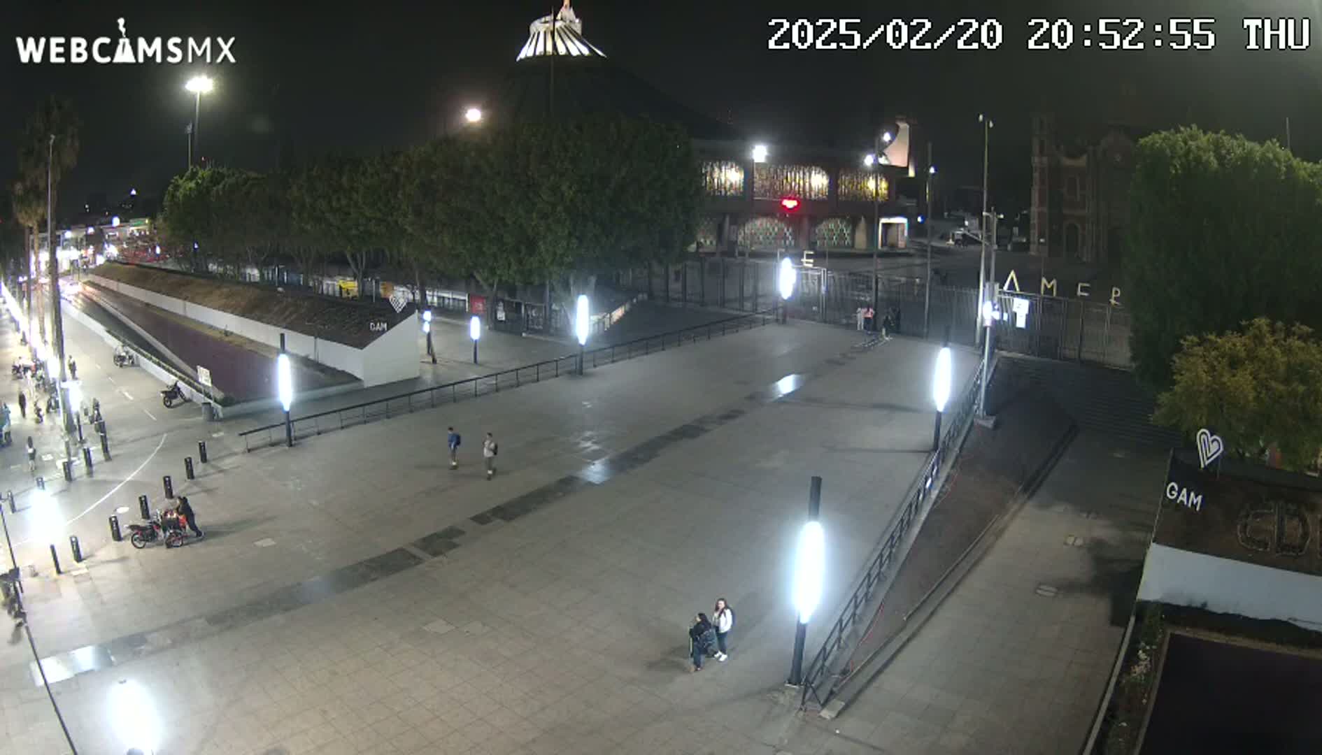 A mostly empty plaza at night is illuminated by numerous bright lights, with a few people scattered about and buildings in the background.