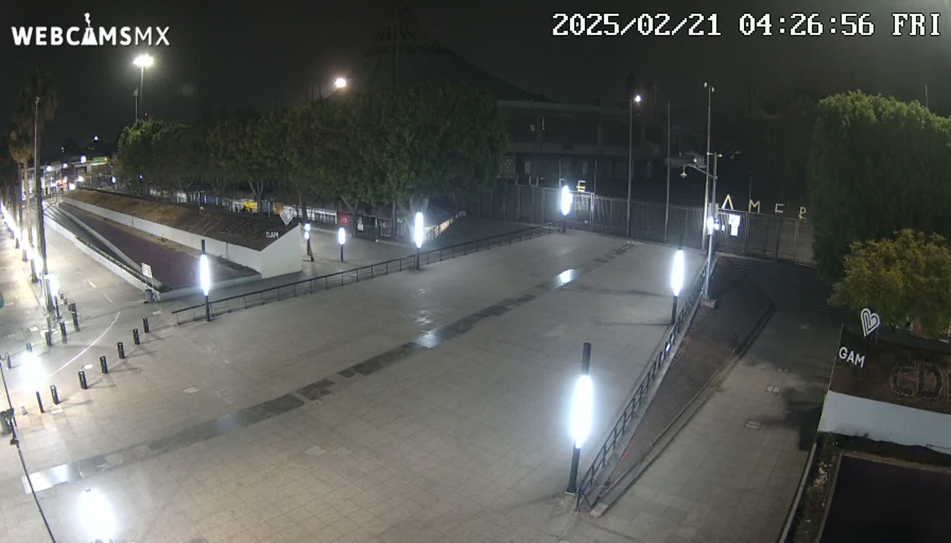 An empty plaza at night, illuminated by streetlights, is bordered by trees and a gated area.