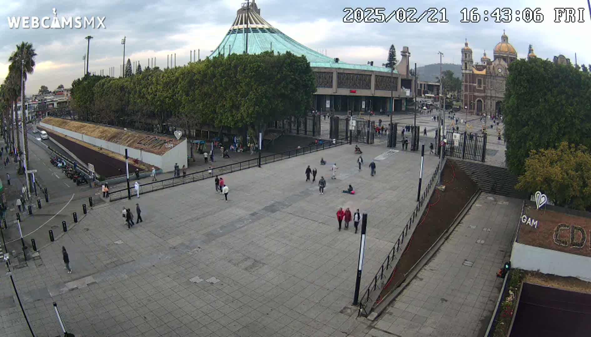 A large plaza with several people walking around, in front of a basilica and a modern building, under a cloudy sky.