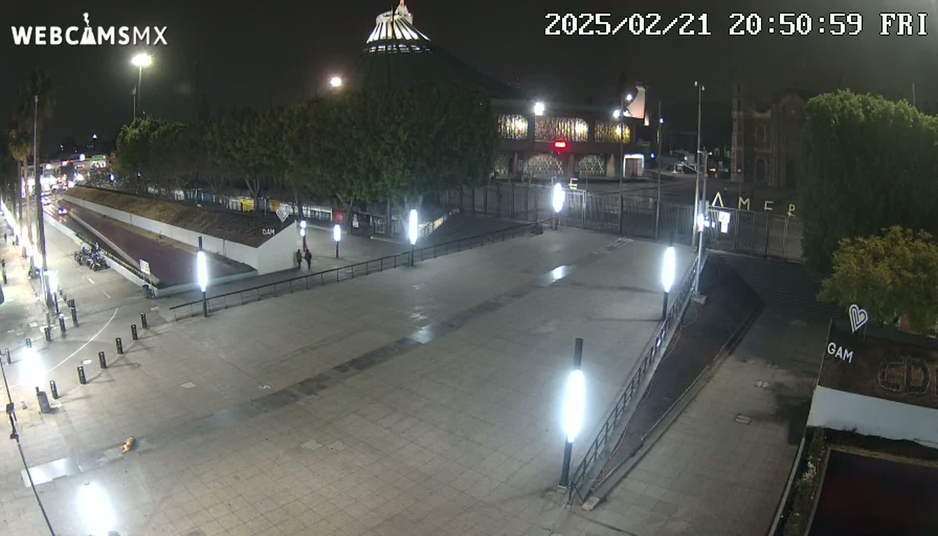 A nighttime view of an empty plaza with several tall lights illuminating a paved area in front of a building.