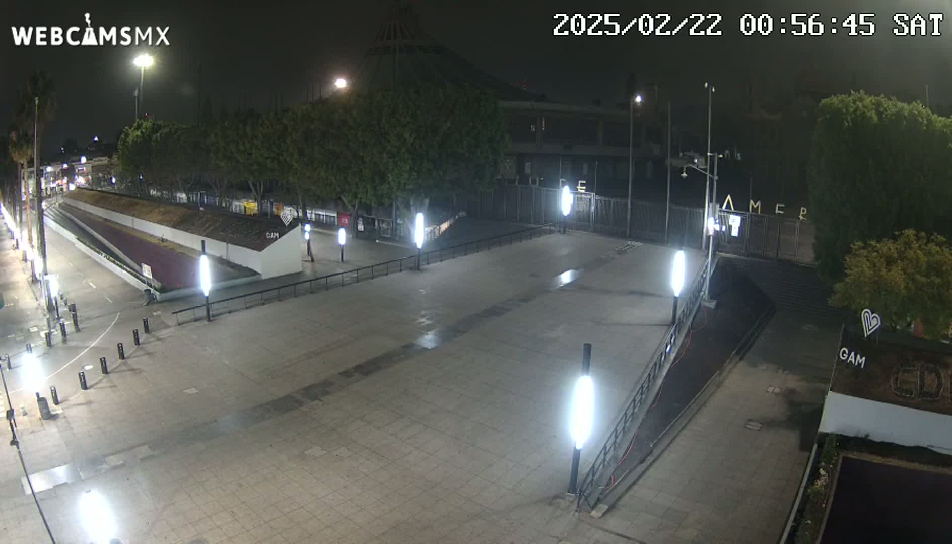 A nighttime view of an empty plaza with several lit lamps, surrounded by trees and buildings.