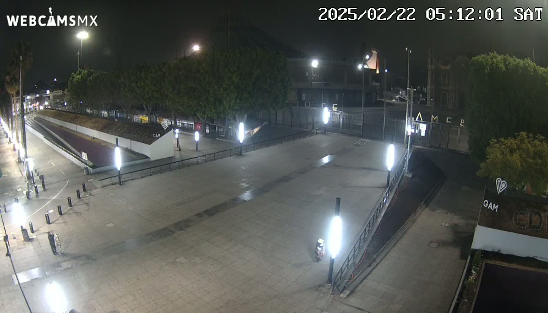 A mostly empty plaza at night, illuminated by numerous lights, is bordered by trees and buildings.