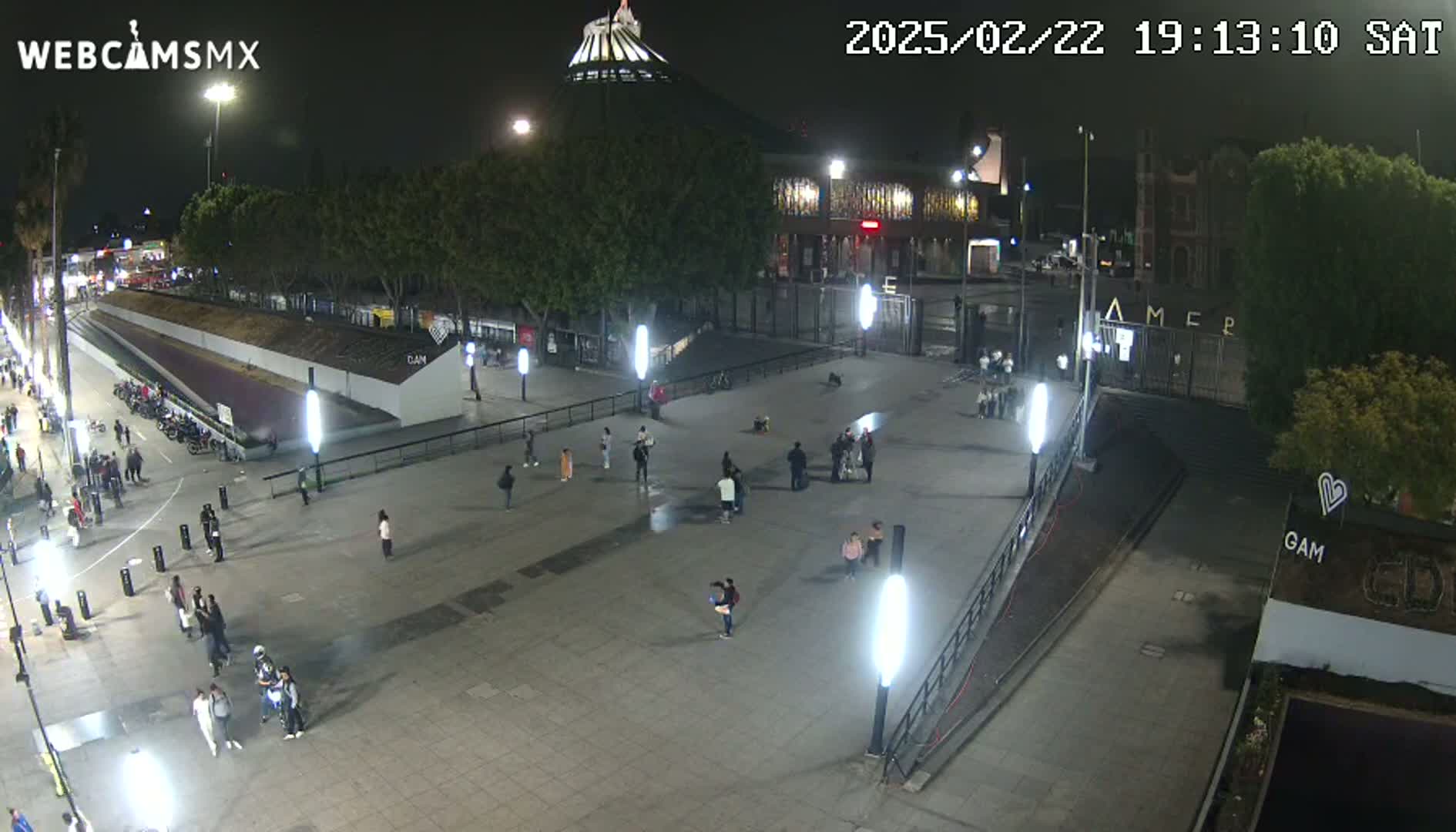 A nighttime, outdoor view of a plaza with people walking around, illuminated by bright lights, and buildings in the background.