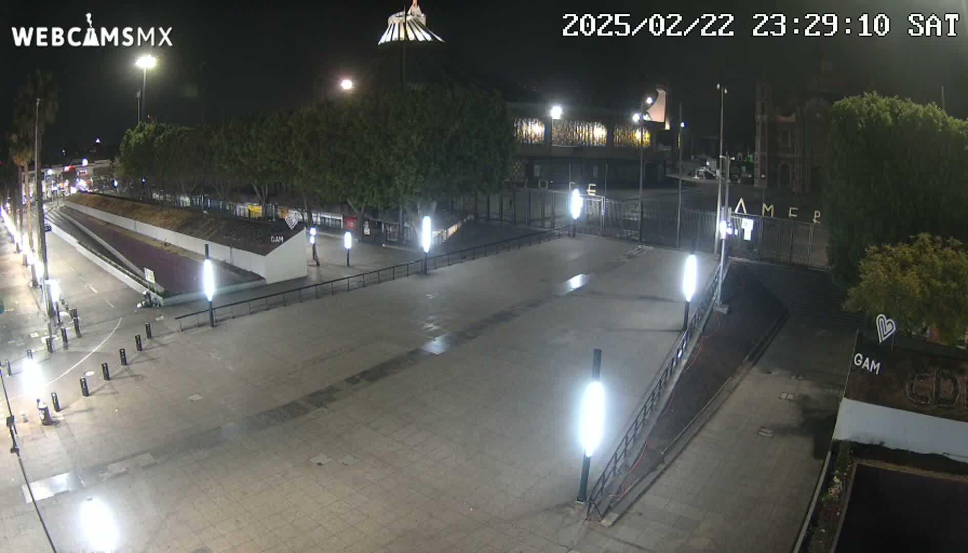 A nighttime view of an empty plaza with buildings and trees in the background, illuminated by streetlights.