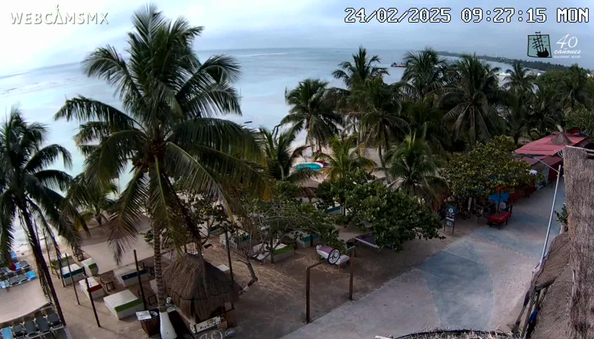 An aerial view shows a beachside area with palm trees, beach furniture, and a paved walkway, under partly cloudy skies.