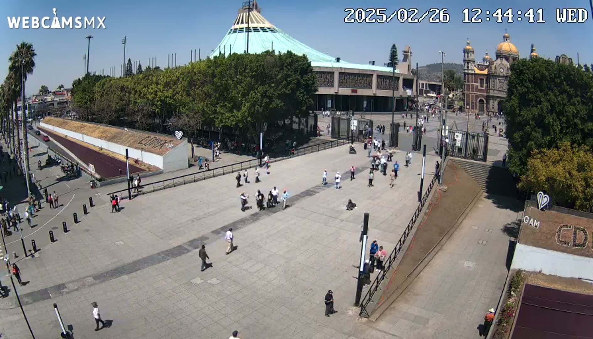 A large plaza with numerous people, flanked by a large basilica and a smaller church, under a clear sunny sky.