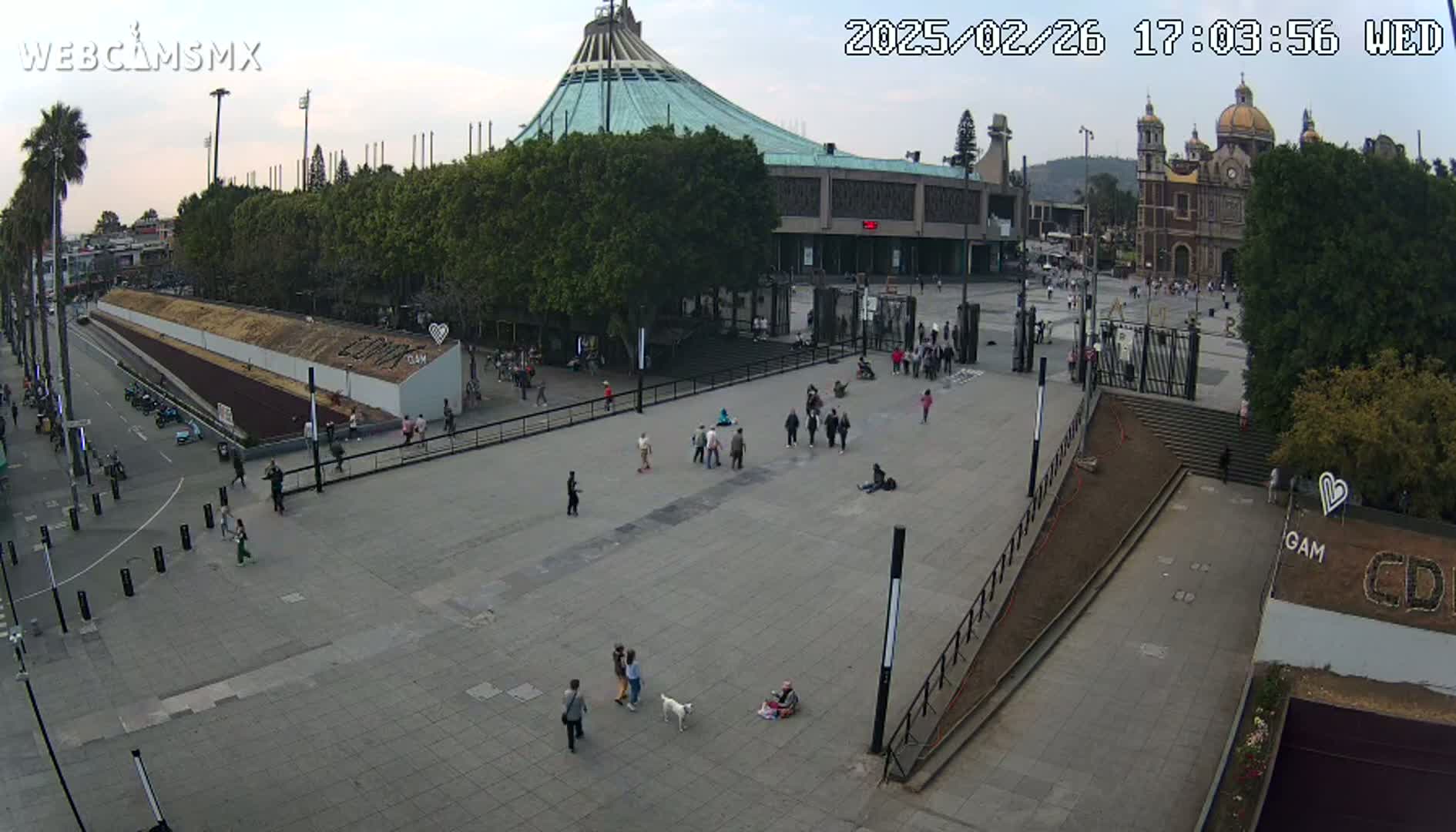 A mostly empty plaza with several people walking around, in front of a large basilica and a modern building, under a partly cloudy sky.