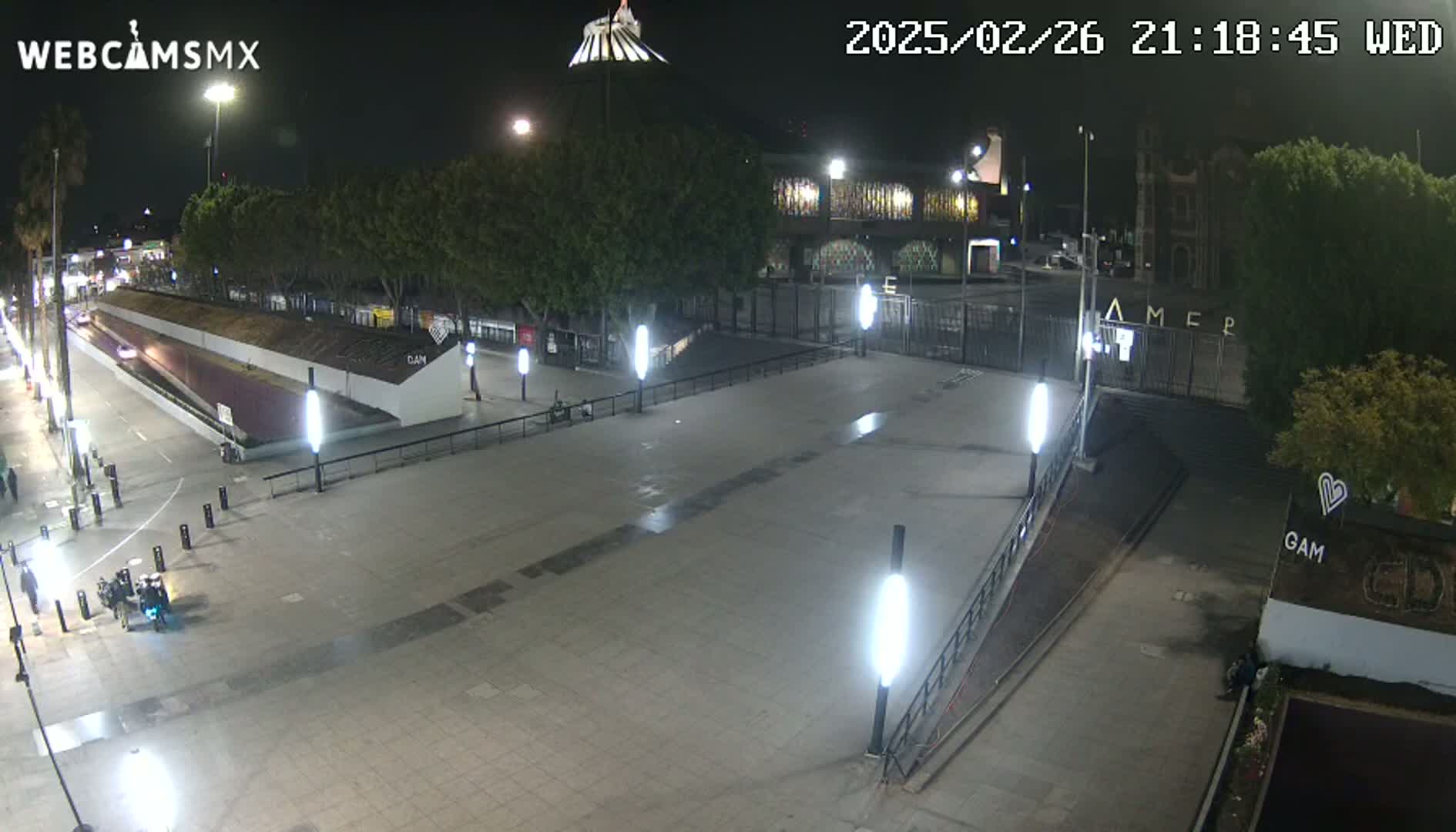 A nighttime, outdoor view of a plaza with several lights illuminating a mostly empty paved area, surrounded by buildings and trees.