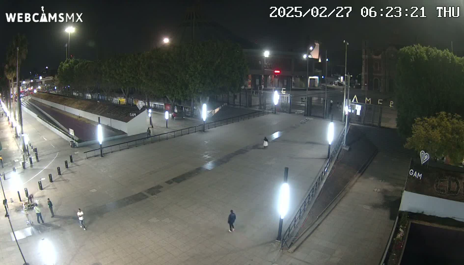 A mostly empty plaza at night is illuminated by numerous bright lights, with a few people visible and trees lining the perimeter.