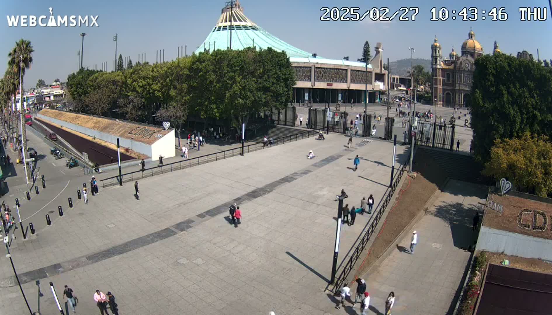 A large plaza with a few people walking around, situated in front of a large basilica and a modern building under a sunny sky.