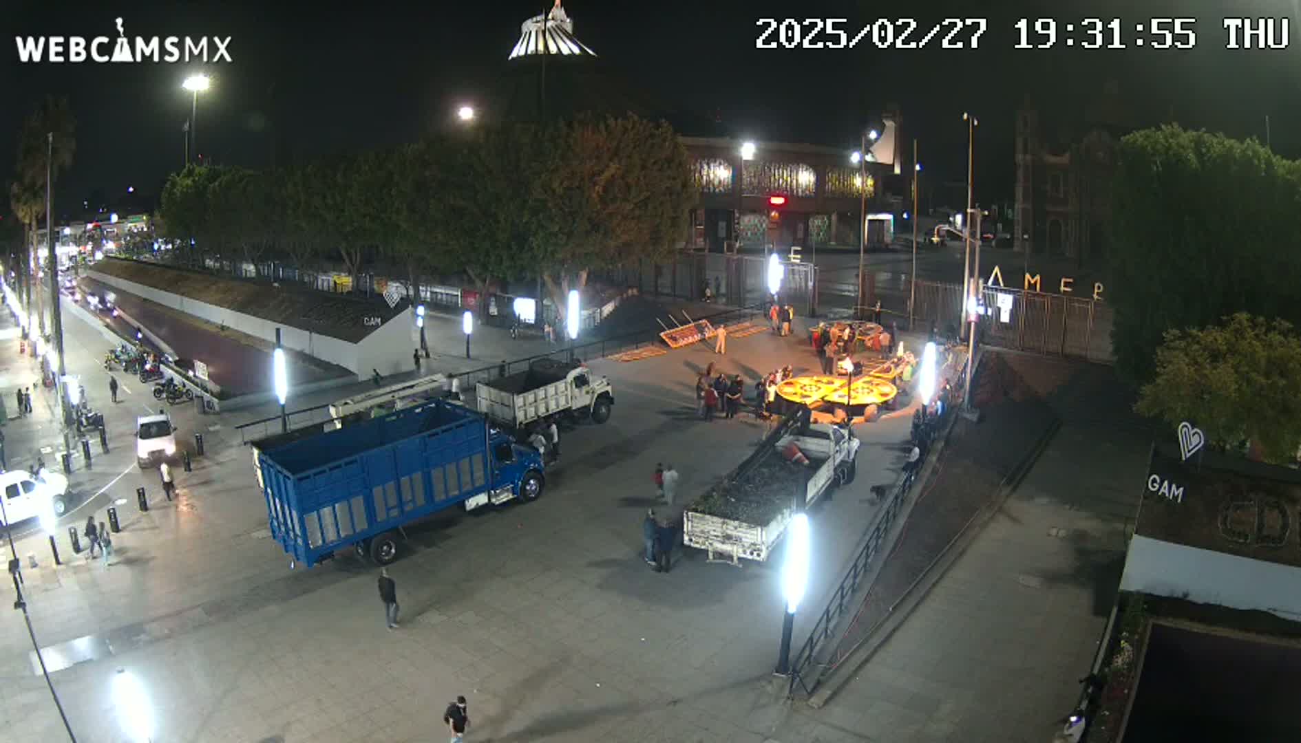 A nighttime aerial view shows a plaza with several trucks, a small group of people gathered around a circular object, and a few pedestrians walking on the surrounding streets.