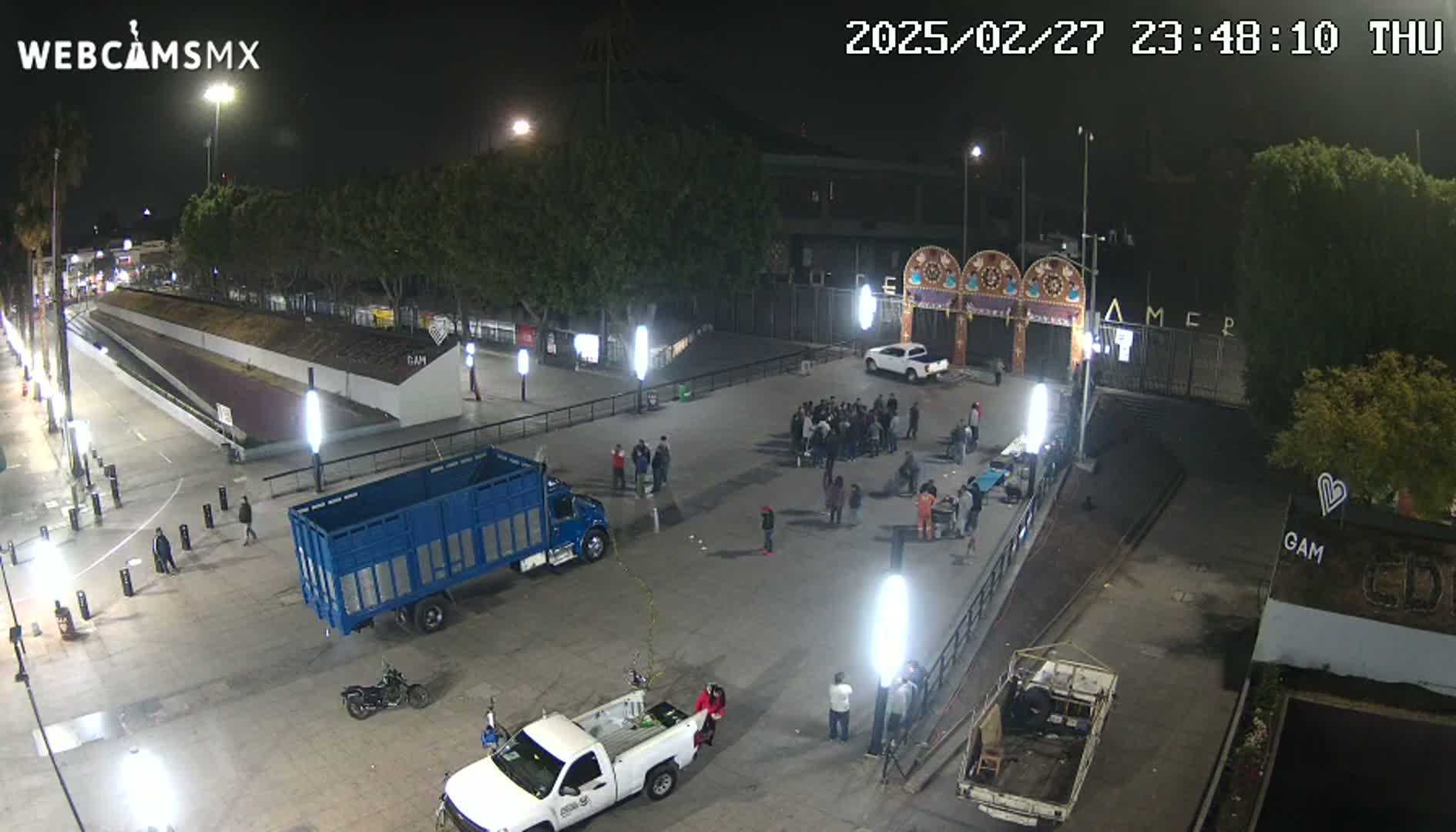 At night, a mostly empty plaza with several vehicles and a group of people gathered together is illuminated by bright lights.
