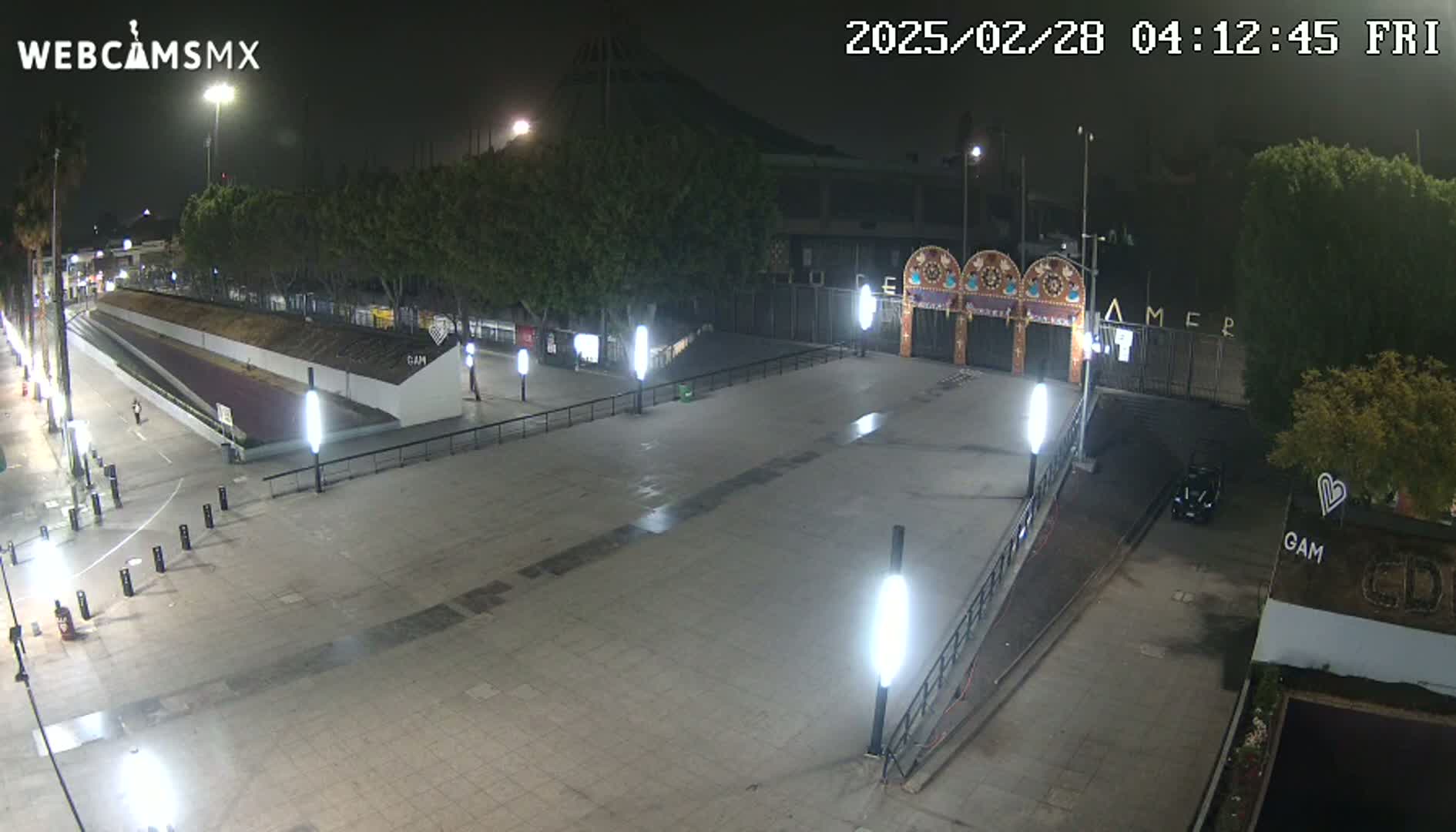 An empty plaza at night, illuminated by streetlights, is situated before a gated entrance adorned with decorative arches.