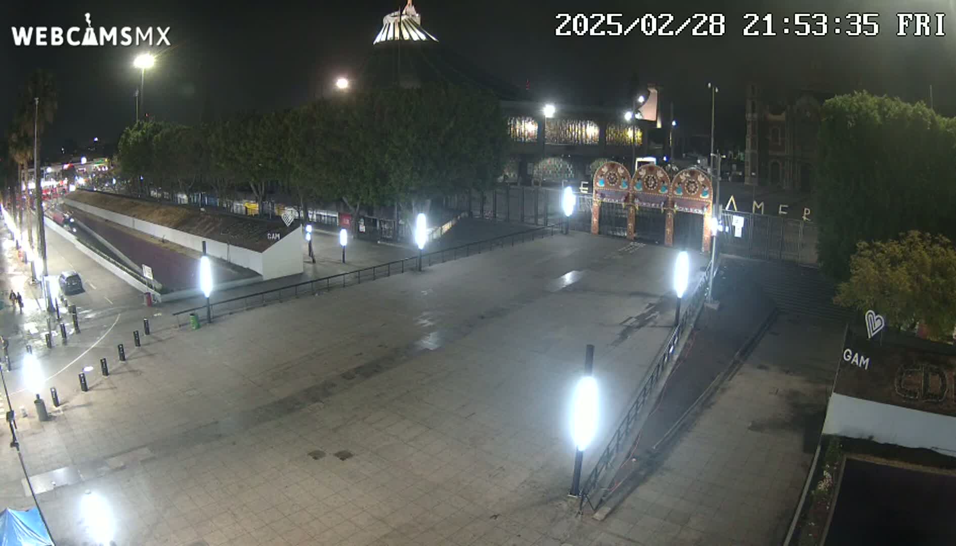 An empty plaza at night, illuminated by streetlights, with a large building and gate in the background under a clear, dark sky.
