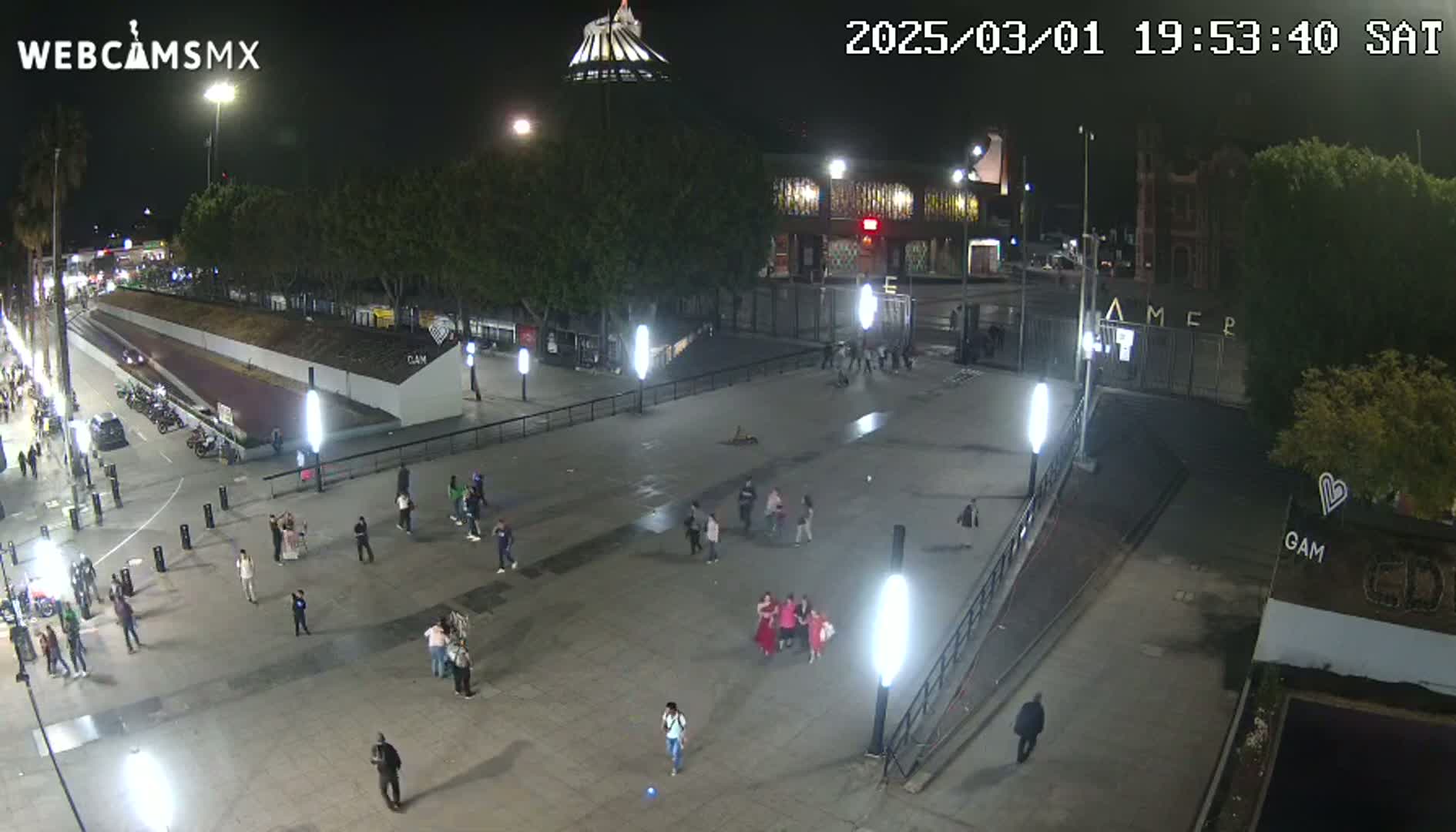A nighttime, outdoor view of a plaza with several people walking around, some buildings, and trees.