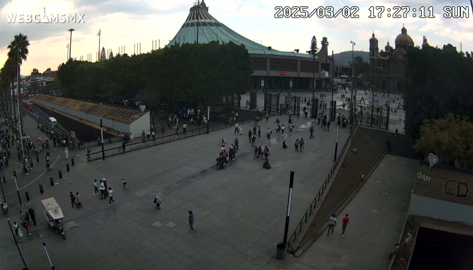 A large plaza with many people, a large basilica, and other buildings is visible under a partly cloudy sky.