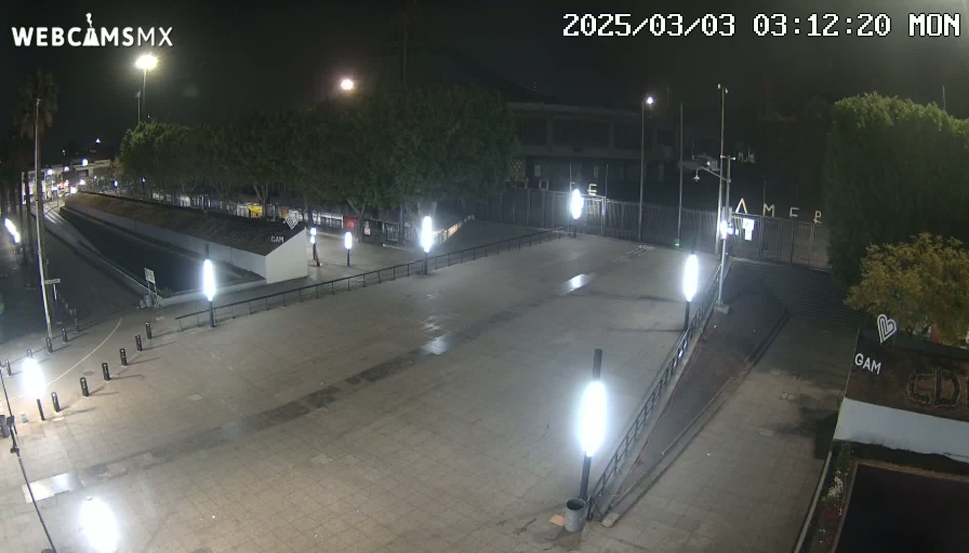 A nighttime, outdoor view of a paved plaza with several lights, trees, and a gated area in the background.
