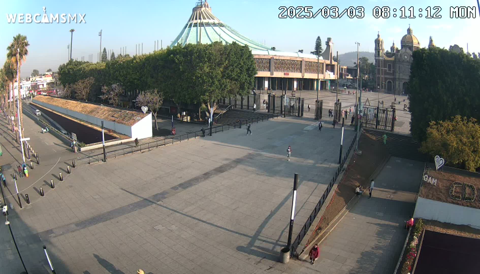 A large plaza with a few pedestrians, in front of a modern basilica and a smaller, traditional church, under a sunny sky.